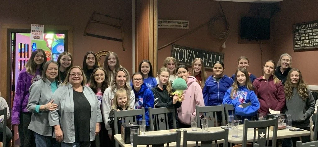 Group of women and girls gathered in a restaurant or cafe, posing for a photo. They are smiling, some wearing casual and others sports hoodies, with a girl holding a plush broccoli toy.