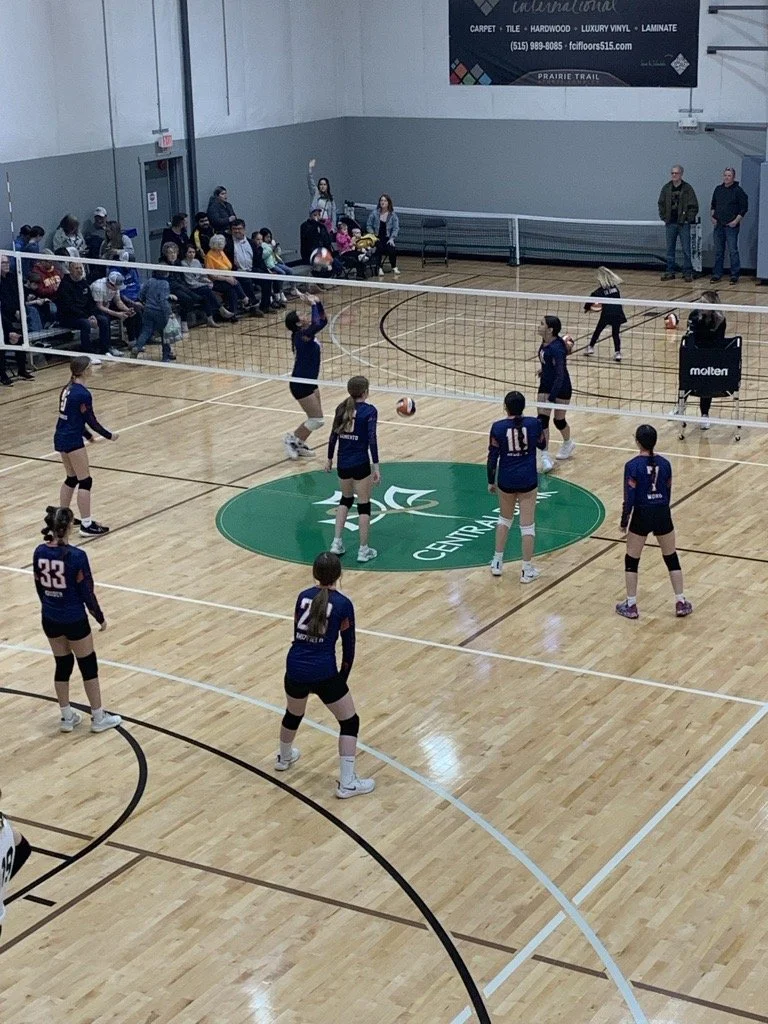 Children playing volleyball on an indoor court with audience seated on benches and standing in the background.
