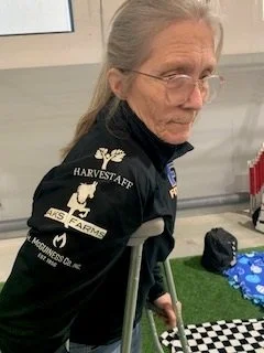 An elderly woman with gray hair, wearing glasses and a black Harvest Staff jacket, leaning on a walker indoors with a black bag and a small blue blanket with white and blue patterns on the floor nearby.