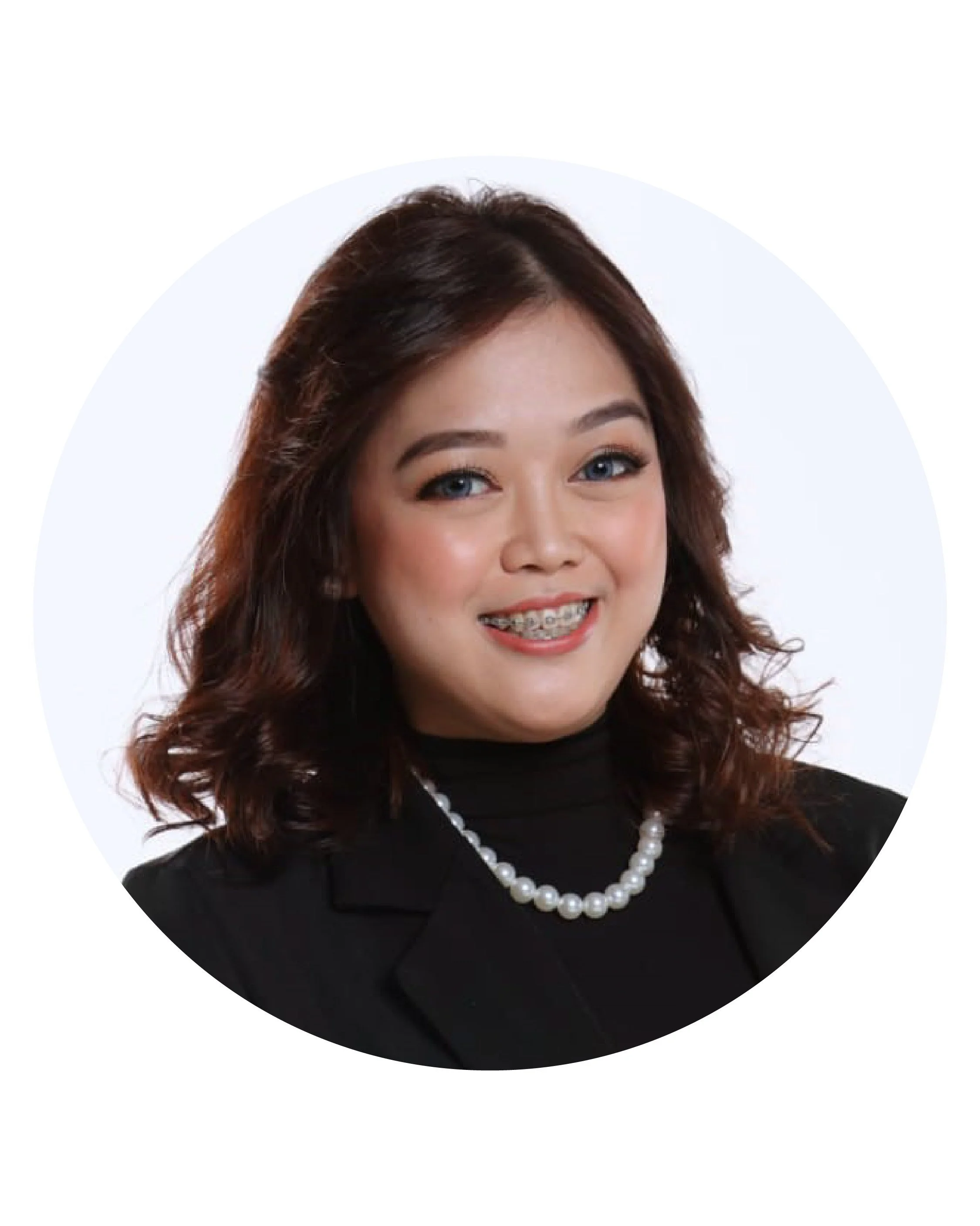 Professional headshot of a woman with shoulder-length wavy brown hair, blue eyes, wearing a black blazer, black turtleneck, and a string of white pearls, smiling with braces, against a plain white background.