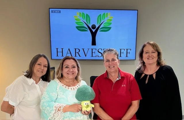 Four women standing in front of a screen displaying a logo with a tree and the words 'Harvest Off'.