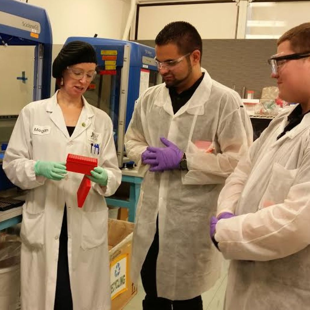 Three scientists in lab coats and safety goggles working together in a laboratory, handling a red object.