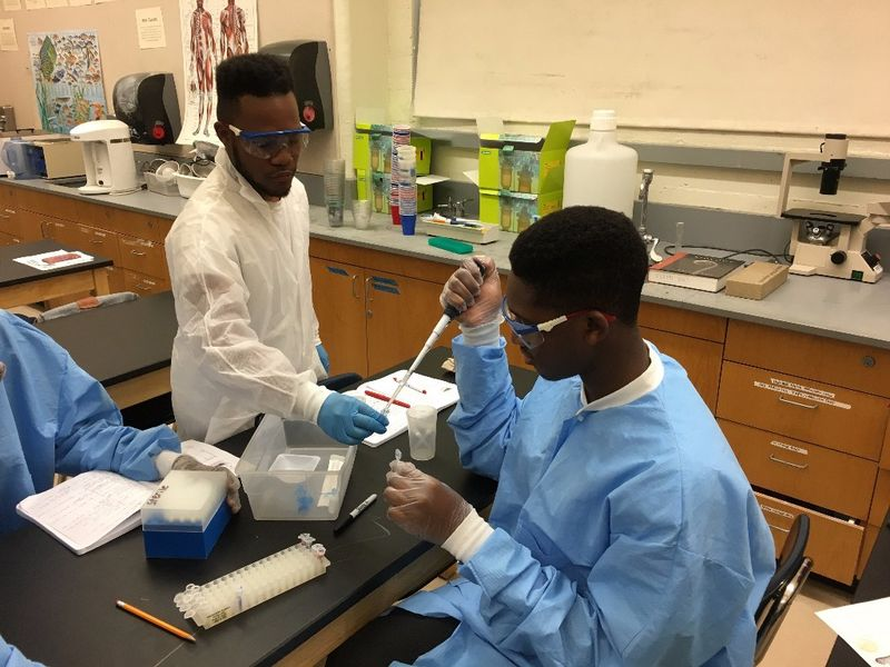 Two students in a science laboratory working with test tubes and laboratory equipment, wearing safety goggles and gloves, with a notebook and scientific supplies on the table.