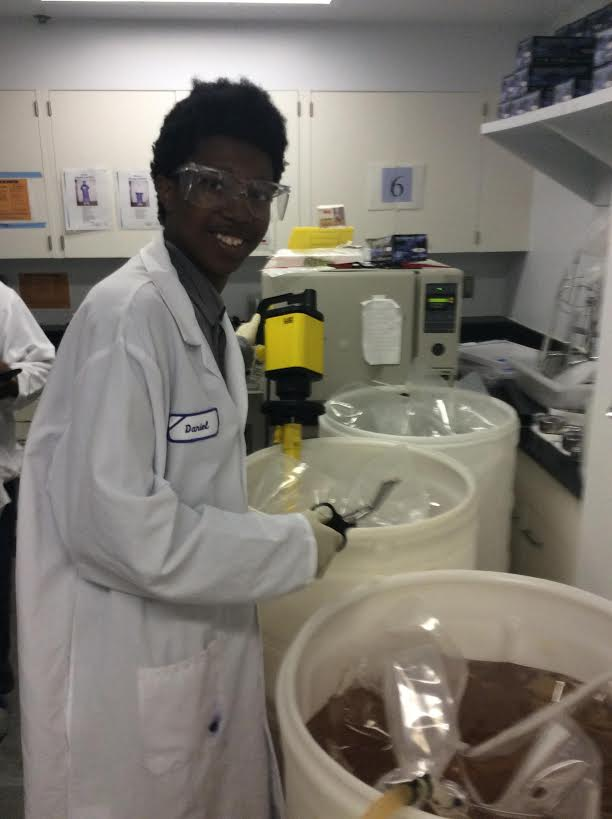 A scientist in a white lab coat and safety glasses working in a laboratory with large containers and scientific equipment.
