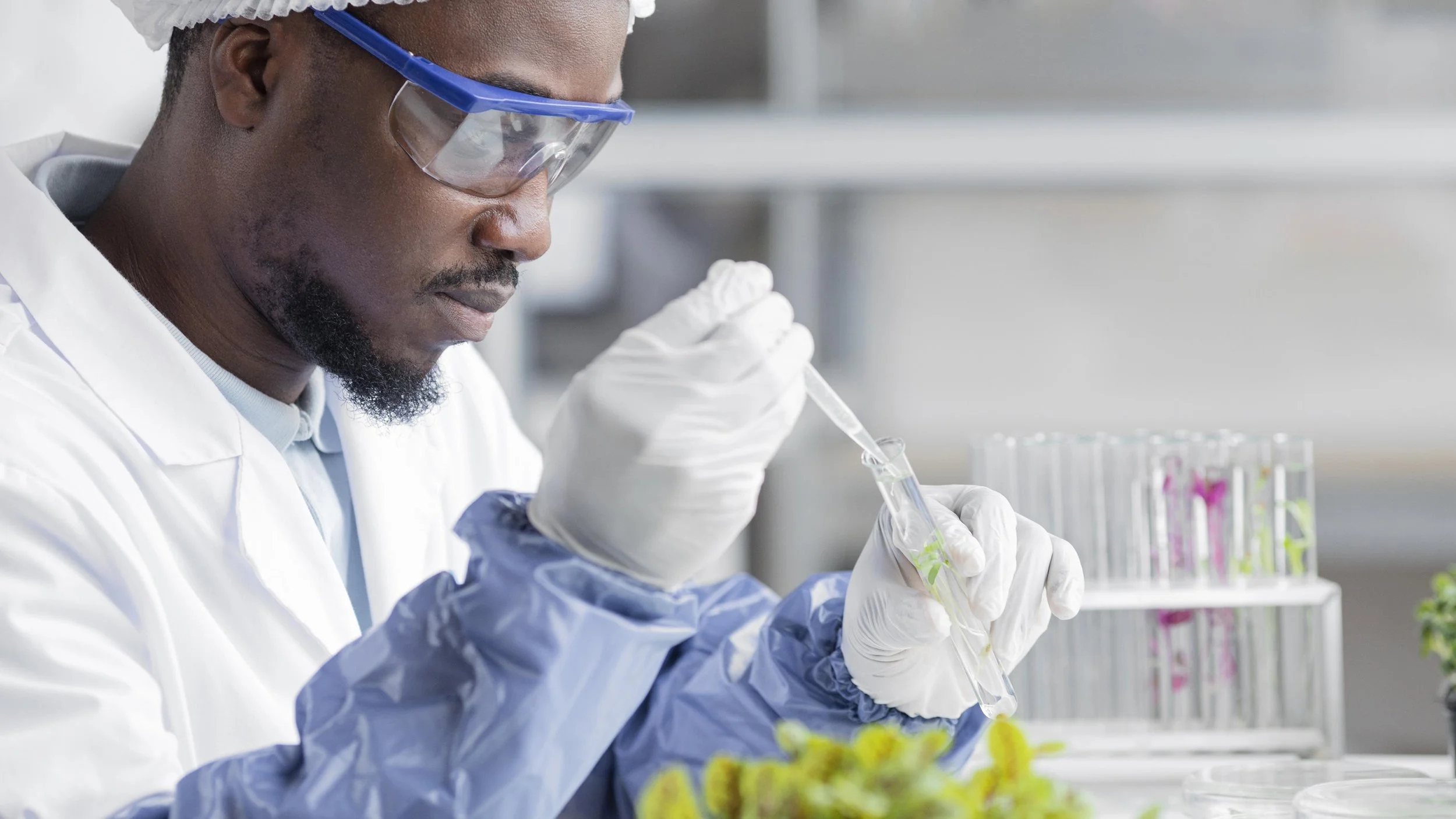 A scientist wearing safety goggles, gloves, and a white lab coat is working with a pipette and test tube in a laboratory with test tubes and plants in the background.