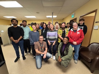 Group of fifteen diverse people posing together inside a room with beige walls and ceiling, some smiling and others with neutral expressions, casually dressed.