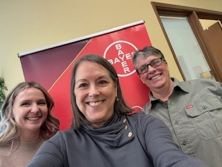 Three smiling people taking a selfie indoors with a Bayer banner in the background.