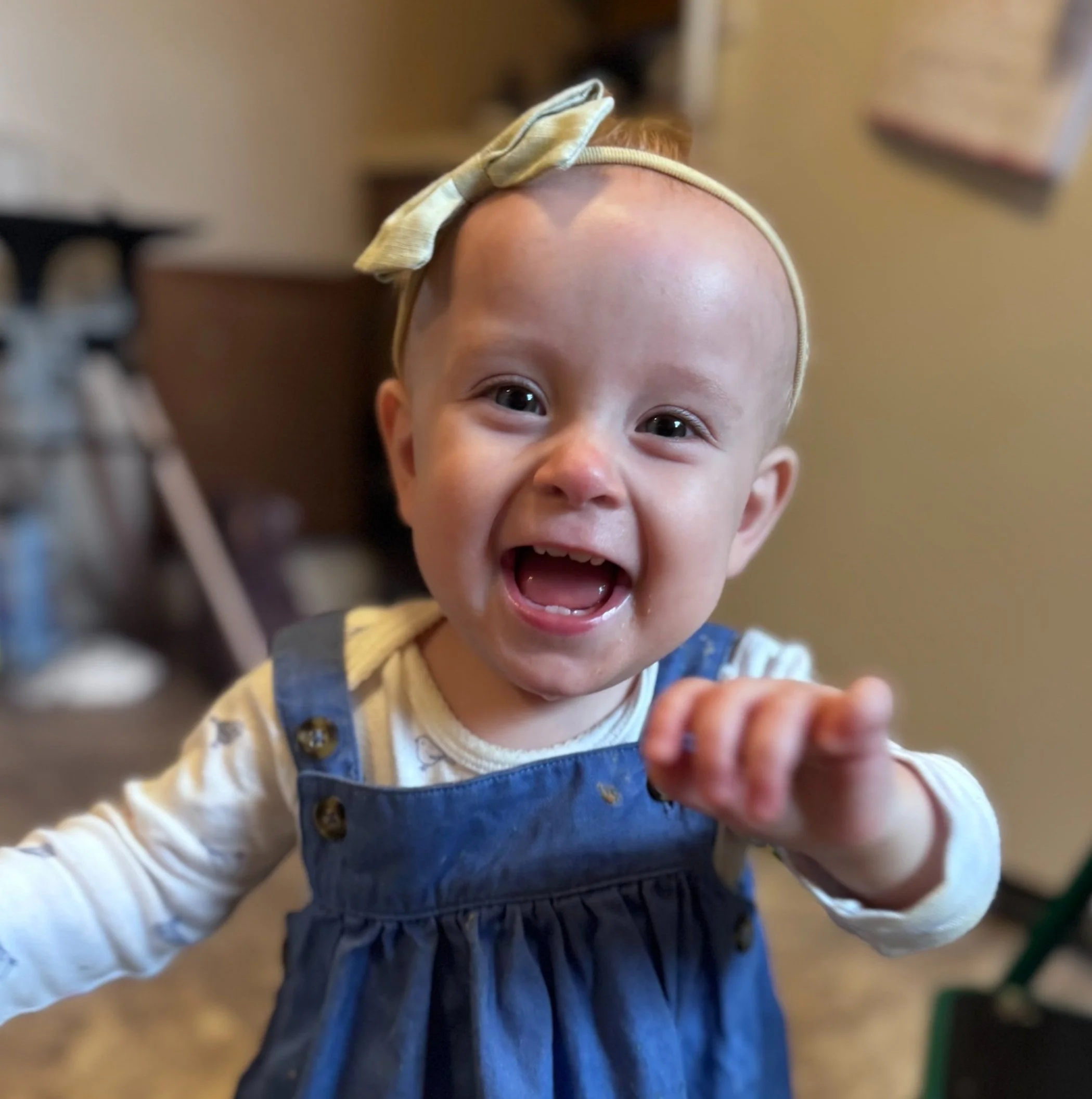 A smiling baby girl with a bow headband, reaching towards the camera, wearing a white shirt and blue dress.