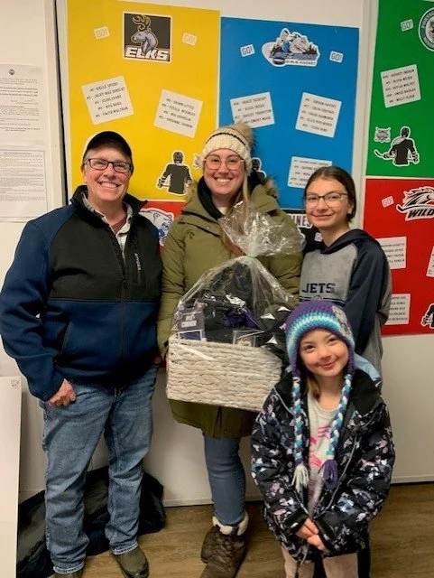 Four people standing indoors, smiling at the camera. Two women and two girls are dressed in casual winter clothing. The woman in the center is holding a gift basket wrapped in clear plastic. Behind them are colorful posters with sports team logos and notes about games.
