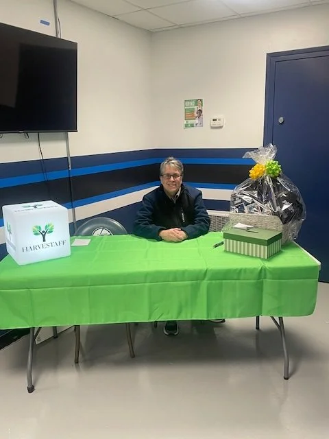 A woman sitting at a table with a green tablecloth, surrounded by gift boxes and a large floral arrangement, in a room with blue and white striped walls.