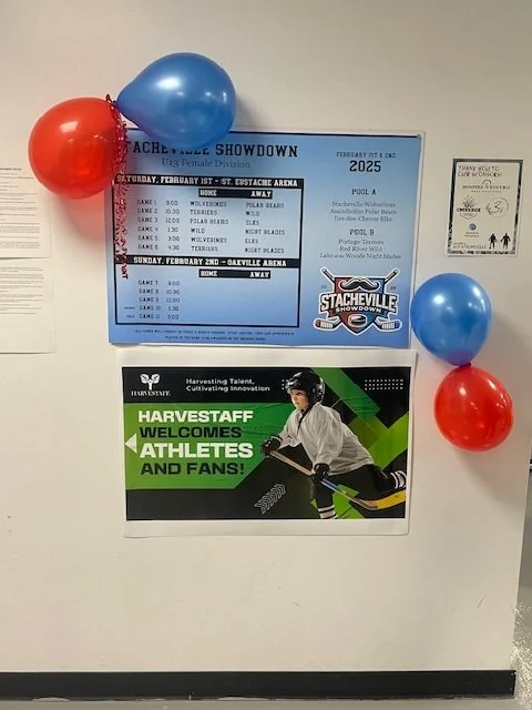 A wall with event posters, balloons, and a small sticker. The larger poster announces a hockey showcase event on February 1st and 2nd, 2025, at the Estherville Arena, featuring teams like Wolves, Polar Bears, and Ice Blades. The smaller poster invites athletes and fans to Harveststaff from Harvesting Talem, with an image of a hockey player in uniform.