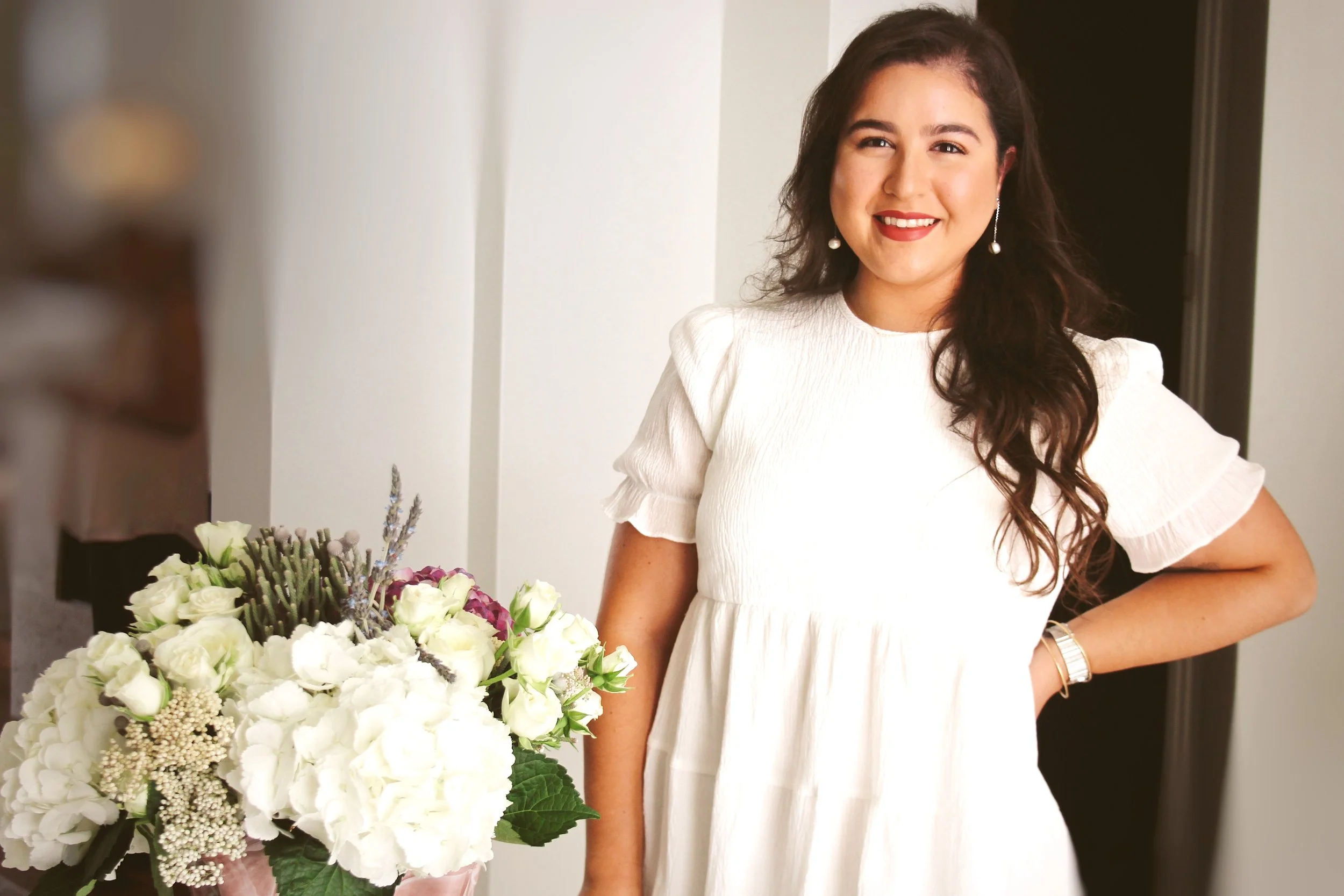 A young woman with long brown hair, wearing a white dress with puffed sleeves and pearl earrings, smiling, standing next to a large bouquet of white and purple flowers.