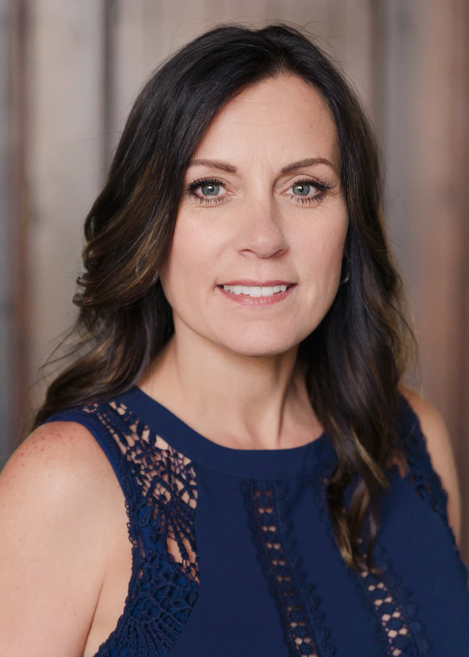 A woman with long wavy dark brown hair and blue eyes wearing a navy blue dress with lace details on the shoulder smiling at the camera.