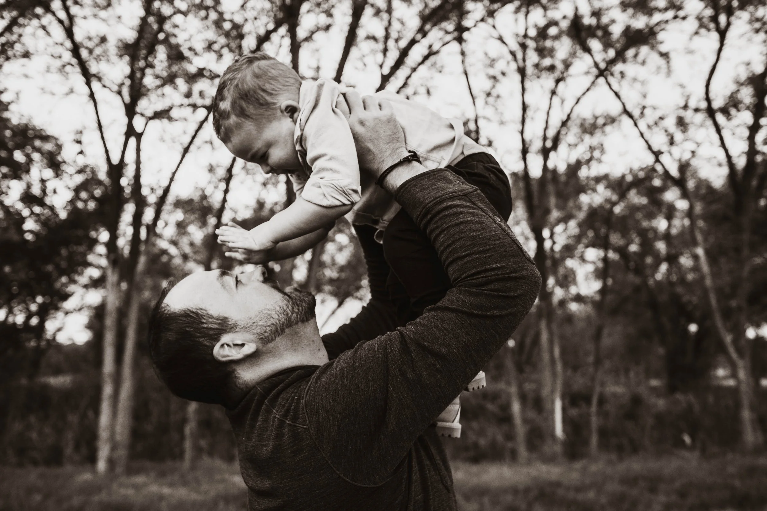 A man lifting a young boy, who is smiling and holding his head, in an outdoor setting with trees in the background.
