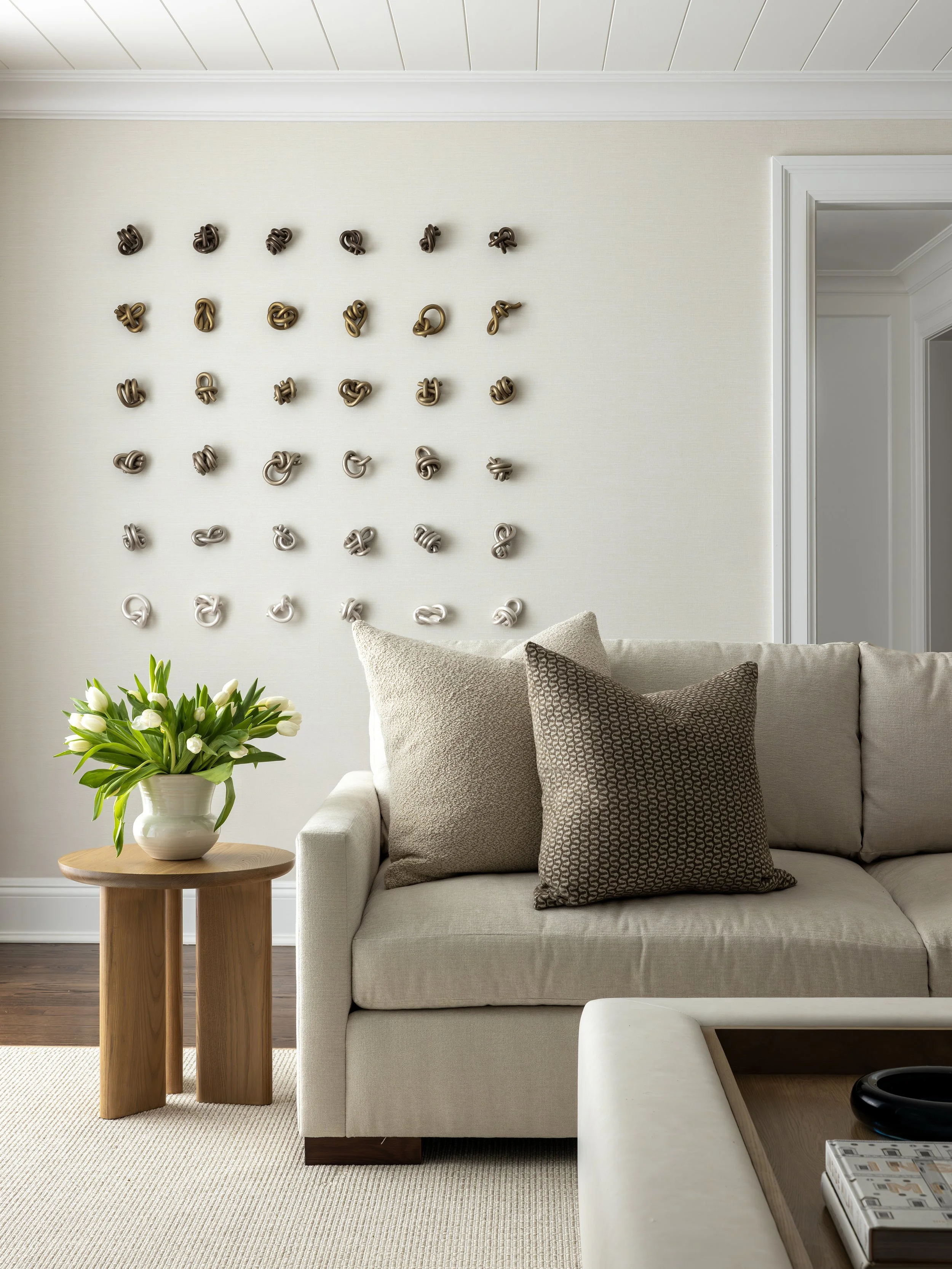 Living room with a beige sofa, brown patterned pillows, a side table with a vase of white tulips, and a wall display of 36 knotted rope art pieces in shades of brown, gray, and cream.