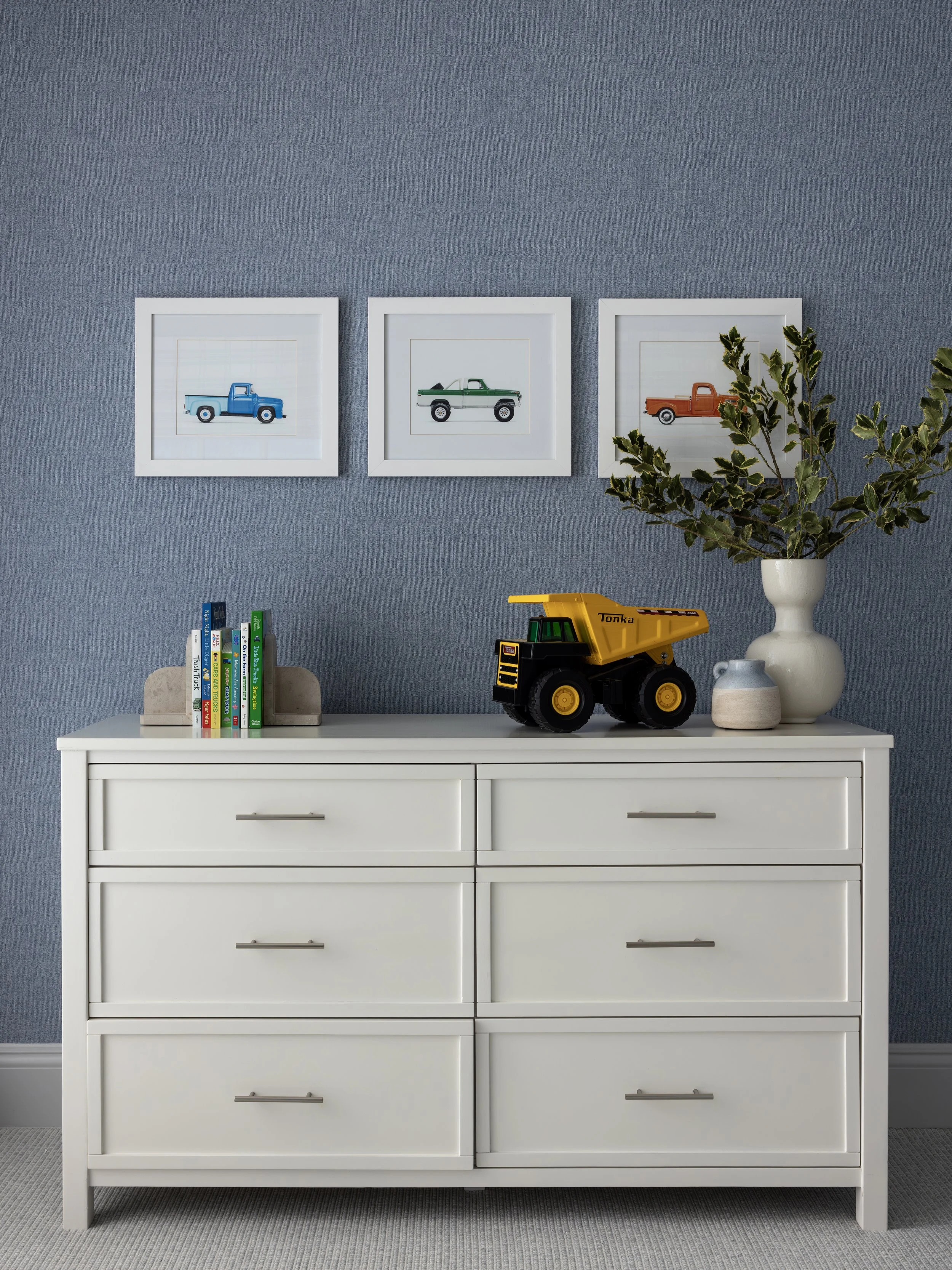A white dresser against a blue wall with three framed pictures of toy trucks, a yellow toy dump truck on top, a cluster of books, and a large white vase with green plant branches.