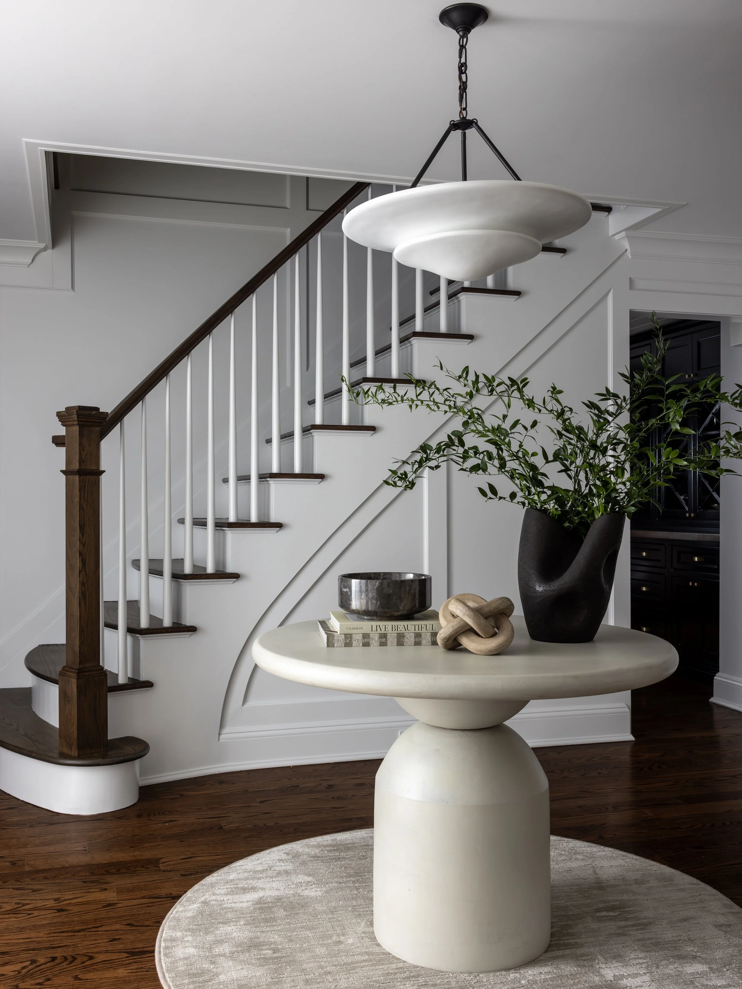 Interior view of a contemporary living space featuring a staircase with white banisters, dark wood steps, and a custom white curved wall. A large black vase with greenery, a stack of books, and decorative objects sit on a unique cream-colored table, 