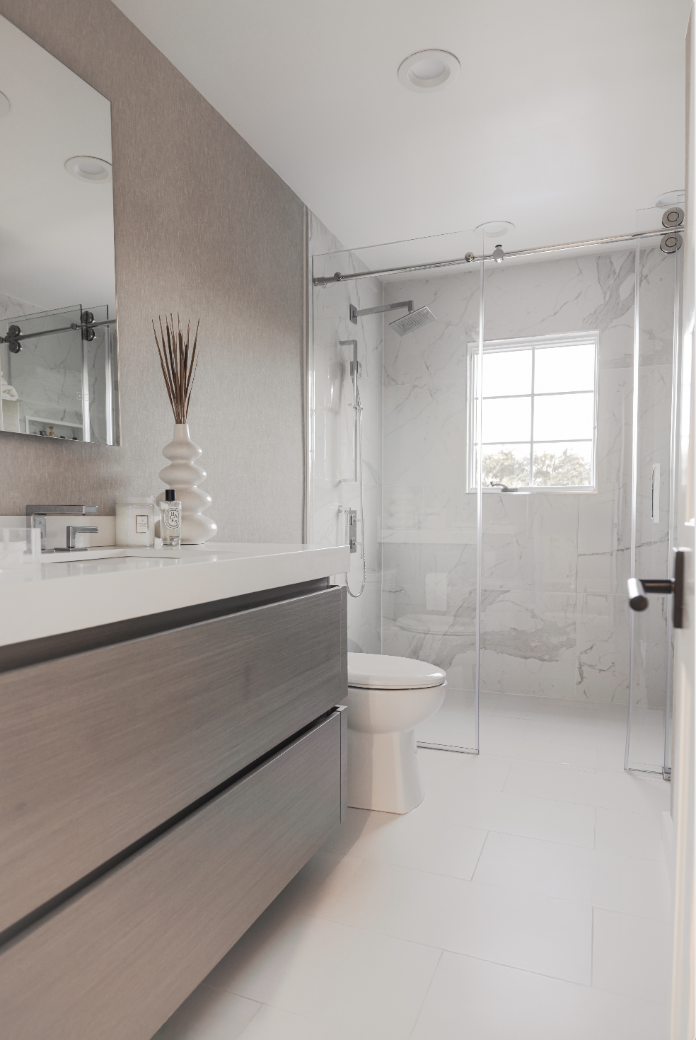 Modern bathroom with a vanity, mirror, and decorative items on the counter. A toilet and a walk-in shower with a glass enclosure, marble walls, and a window letting in natural light.