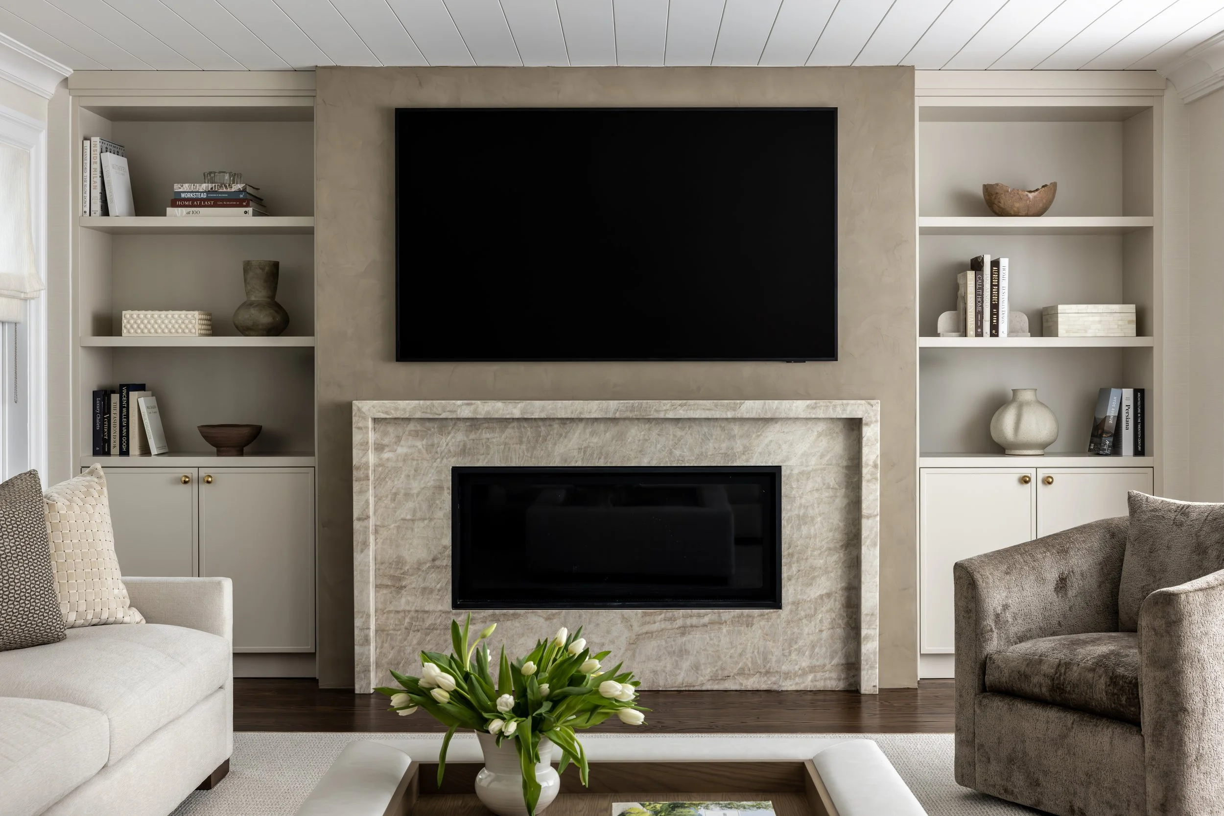 Living room with built-in white shelves, a large TV above a marble fireplace, and a beige armchair. A coffee table with a vase of white tulips is in the foreground.