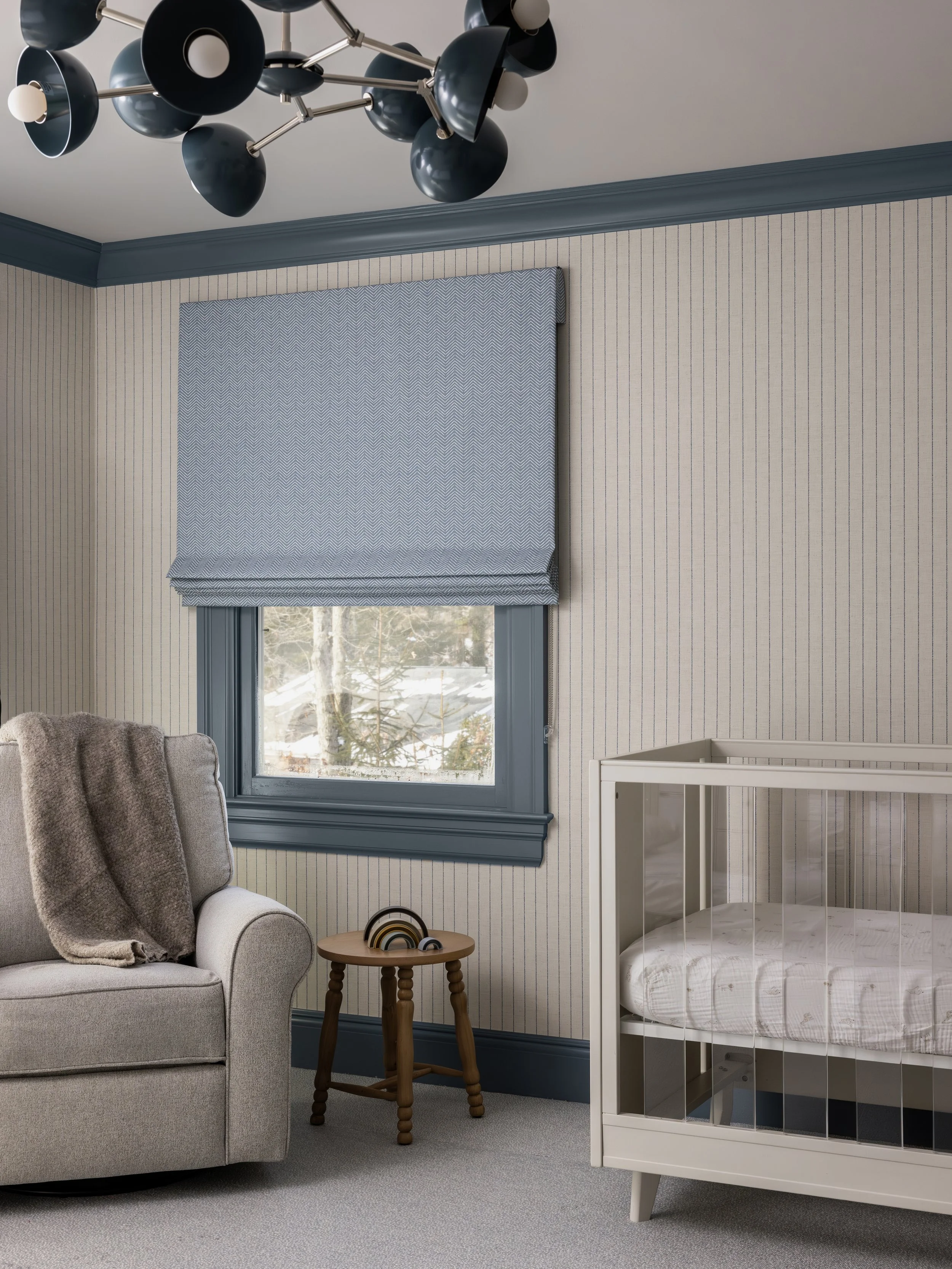 A cozy nursery room with a beige couch, a small wooden side table, a crib, a window with blue trim, a patterned window shade, a modern black and white pendant light, and snow visible outside.