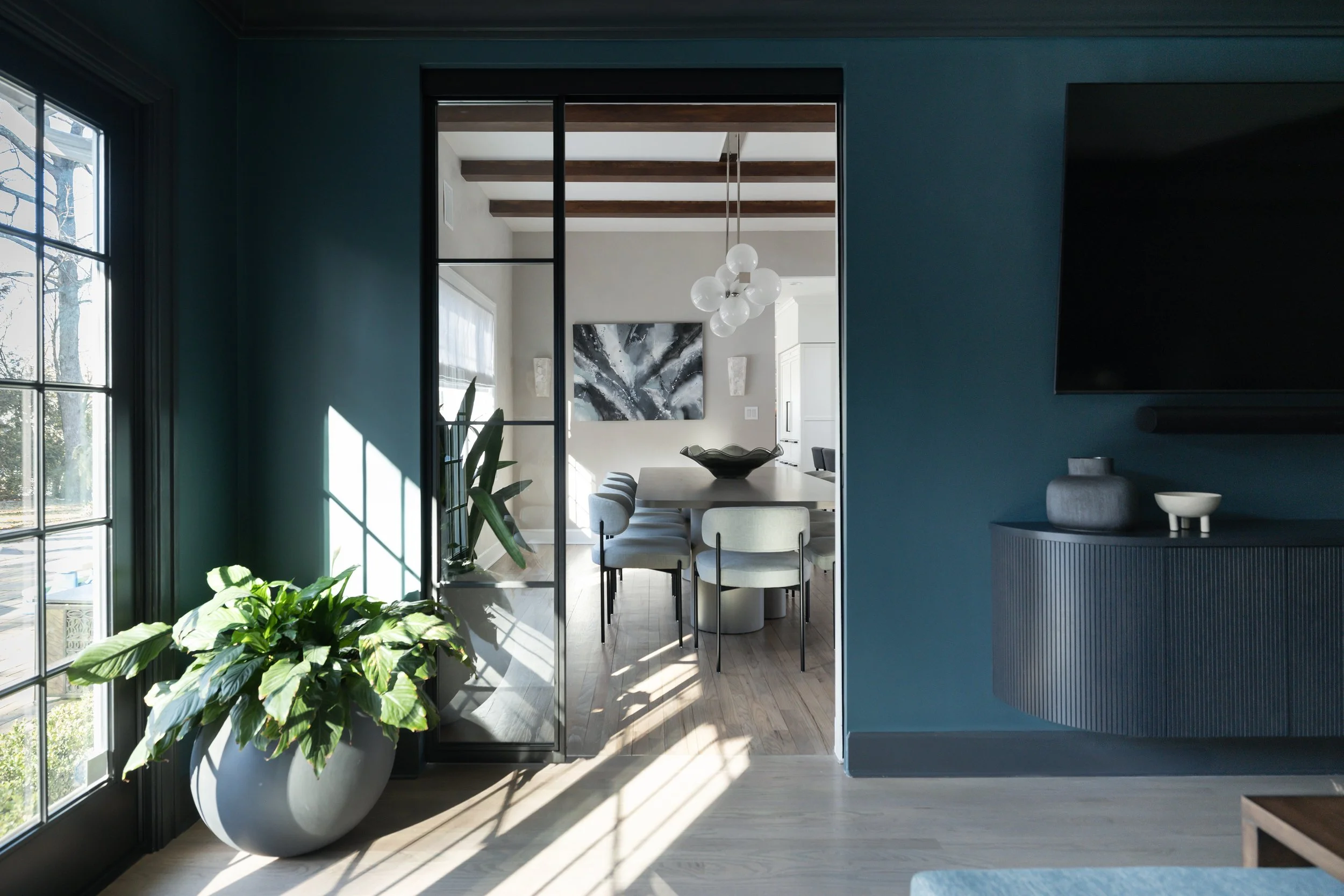 Interior view of a modern dining room through a glass sliding door, showing a wooden table with beige chairs, a black and white abstract painting on the wall, hanging white globe light fixture, and houseplants near the window.