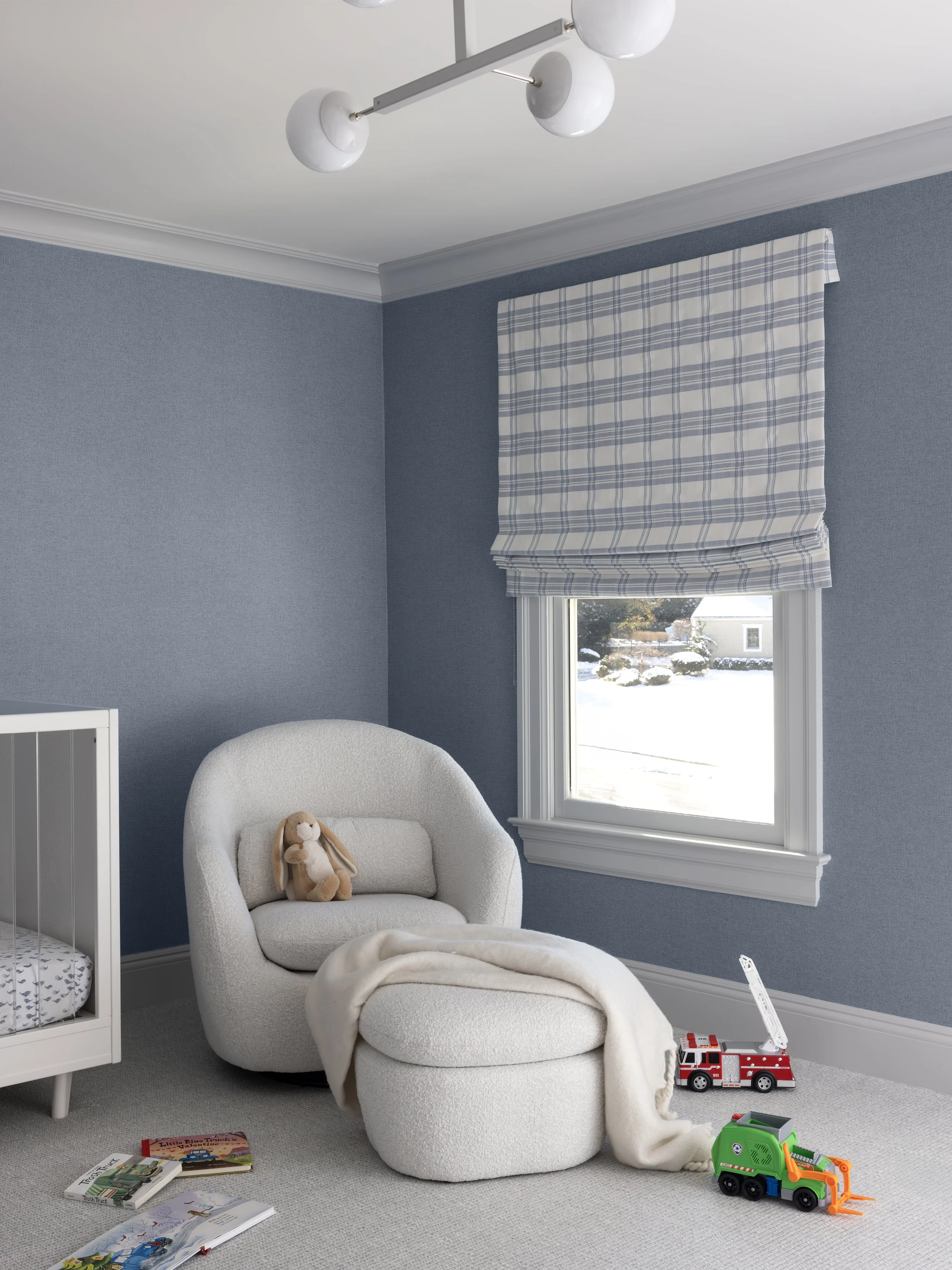 A baby nursery with blue walls, a white armchair with a teddy bear and throw blanket, a window with a checked Roman shade, toys on the floor including a fire truck and a toy garbage truck, and books spread out.