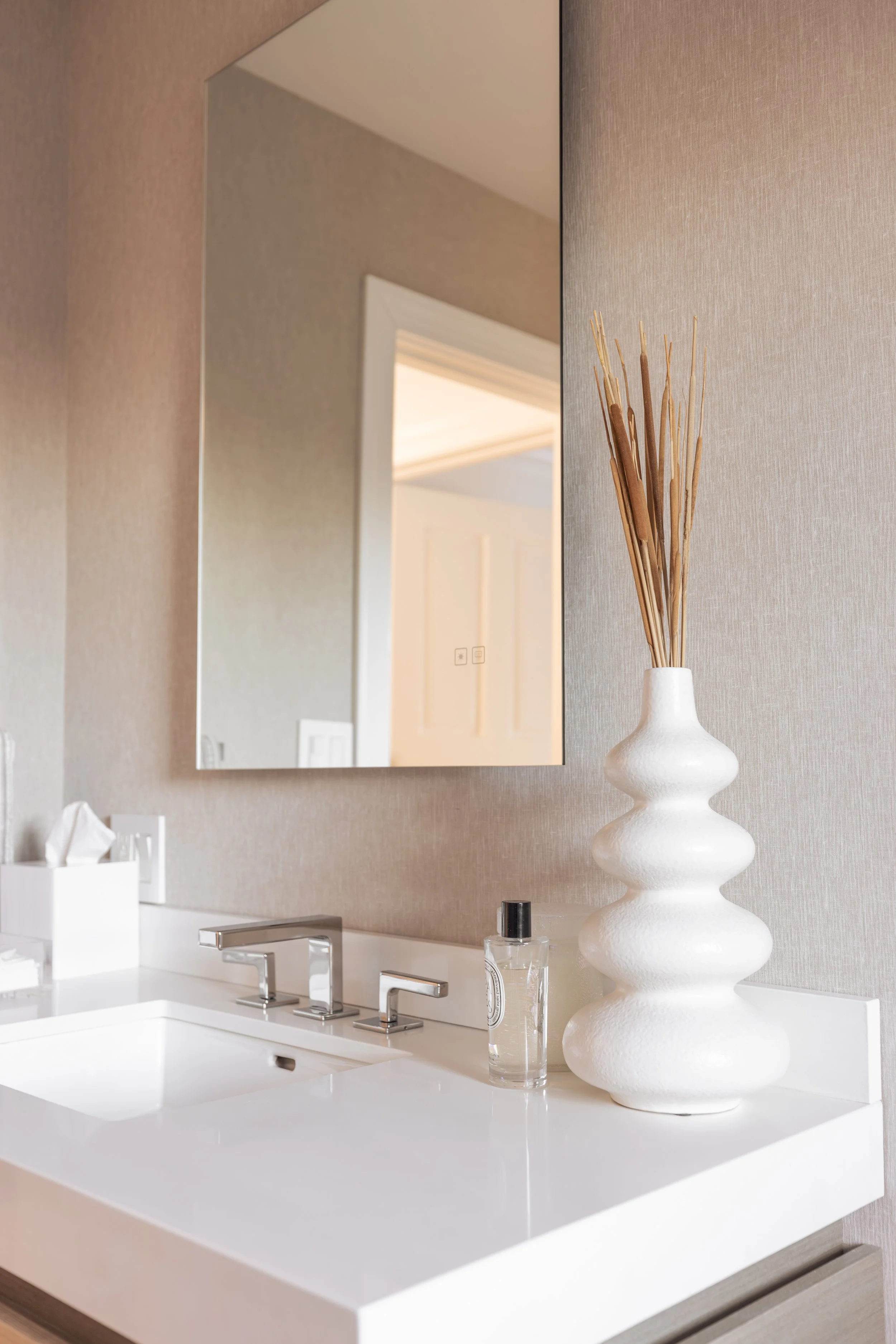 A bathroom countertop with a large white vase holding decorative sticks, a bottle of hand sanitizer, and a rectangular mirror reflecting a door and part of the ceiling.