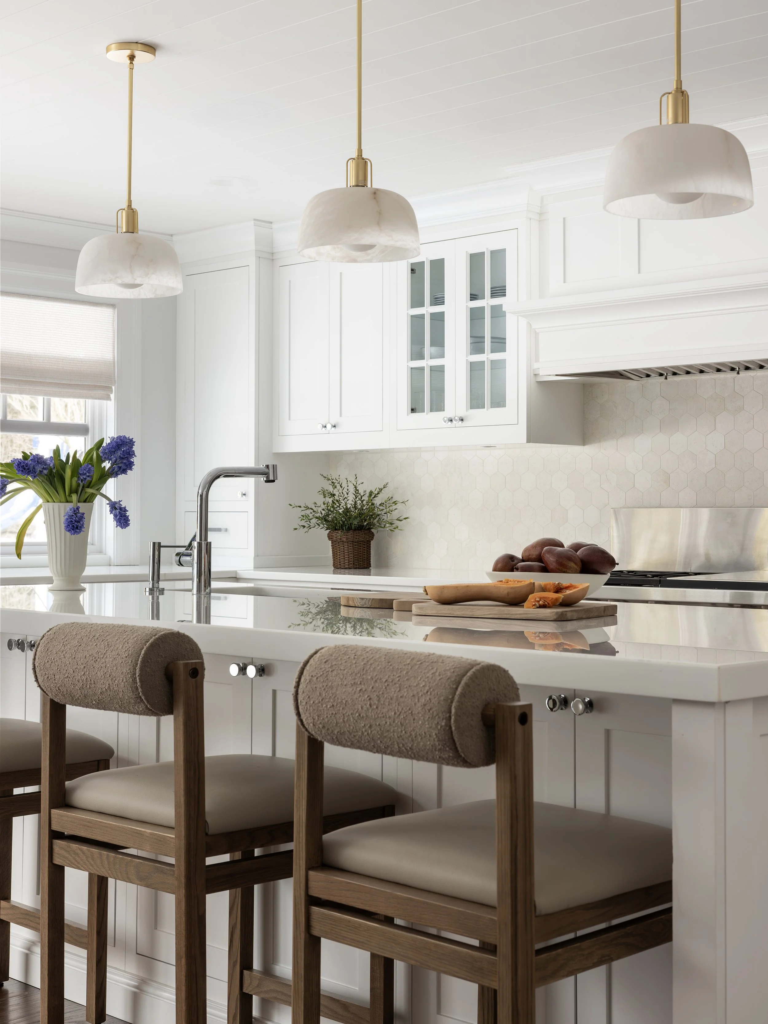A bright, modern kitchen with white cabinetry, a white island with a sink, and three beige bar stools with wooden legs. There are hanging white pendant lights with gold accents. On the countertop, there is a vase of purple flowers, a potted plant, an