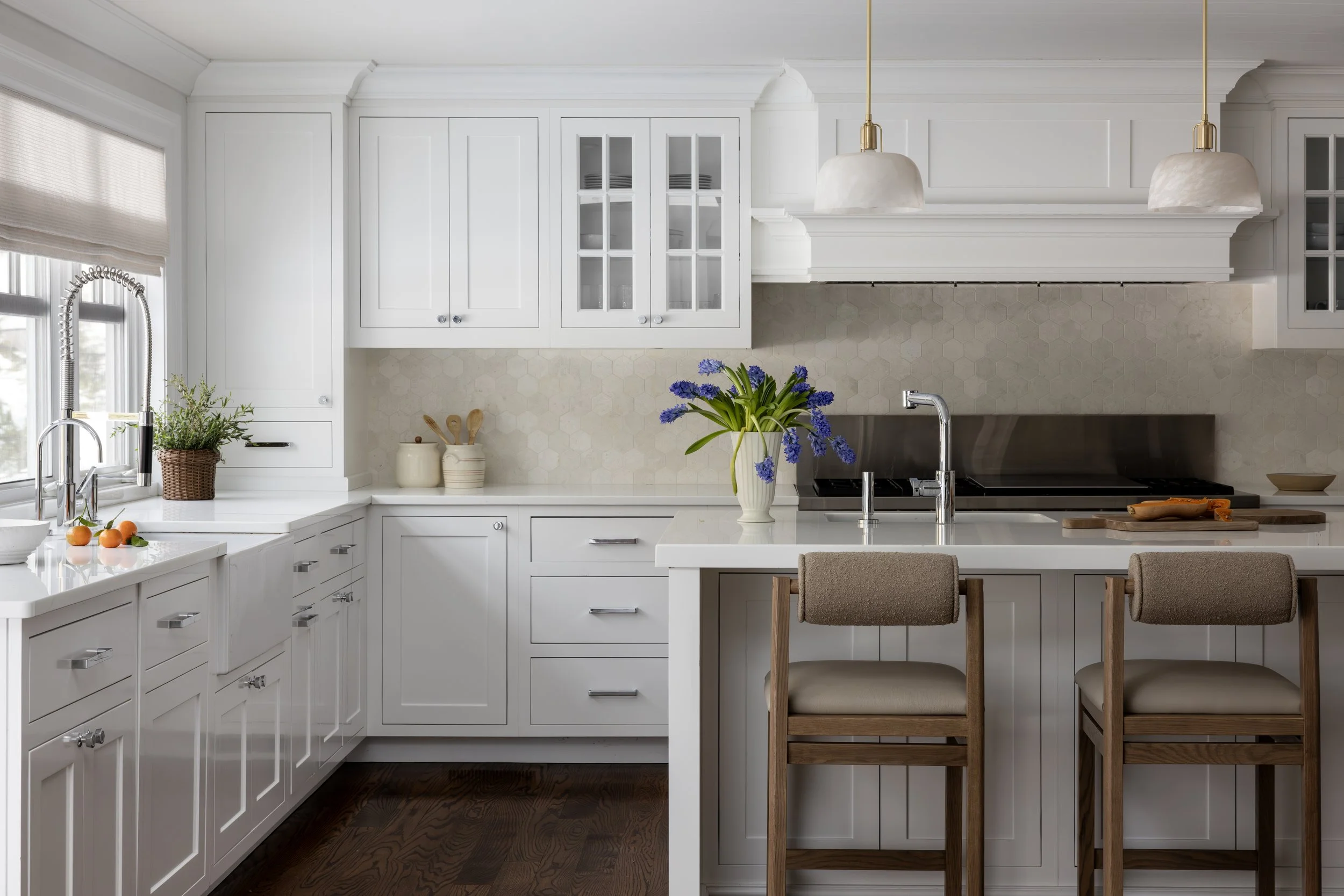 Bright white kitchen with white cabinets, a kitchen island with two beige upholstered bar stools, a vase with purple flowers, and hanging white pendant lights.