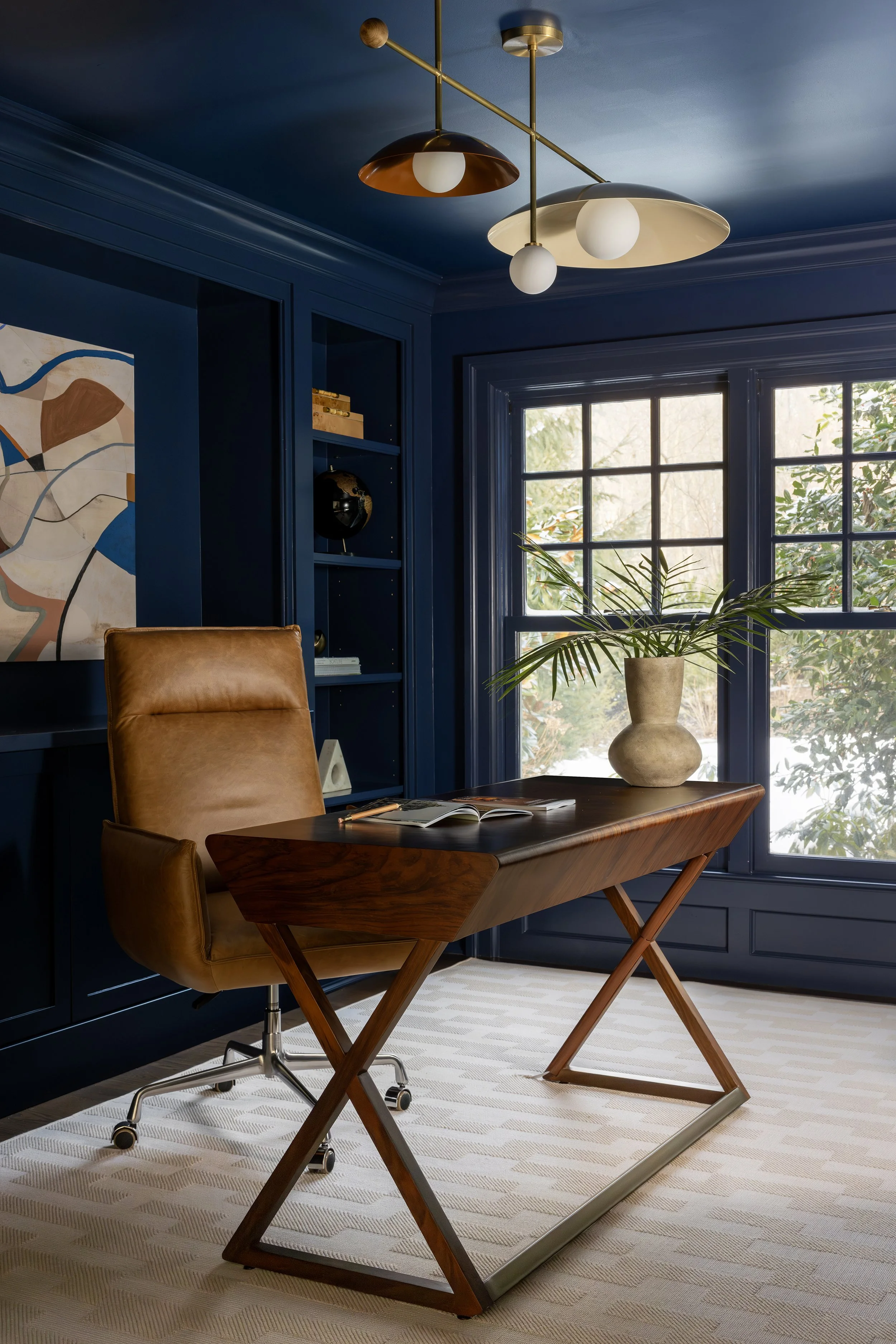 Home office with dark blue walls and ceiling, wooden desk, tan leather office chair, large window with greenery outside, decorative light fixture on ceiling, bookshelf with decorations, and abstract painting on wall.