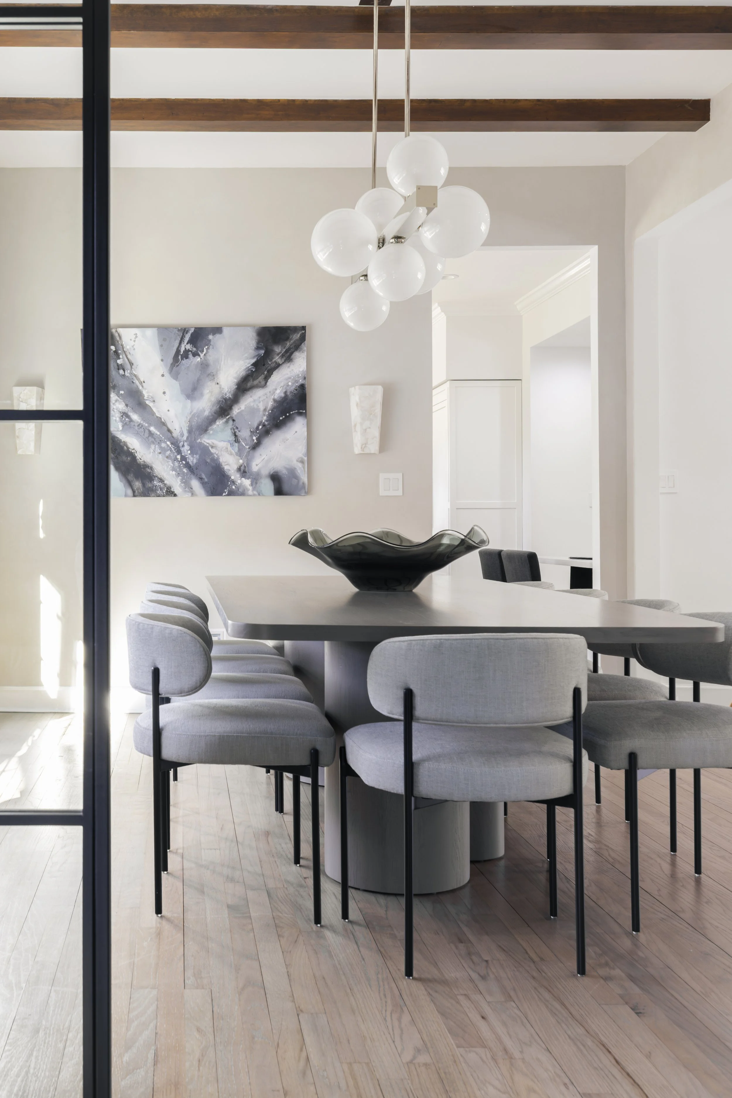 Modern dining room with a large gray table, surrounded by light gray upholstered chairs with black legs, a black decorative bowl on the table, a contemporary chandelier with globes hanging from the ceiling, an abstract black and white painting on the