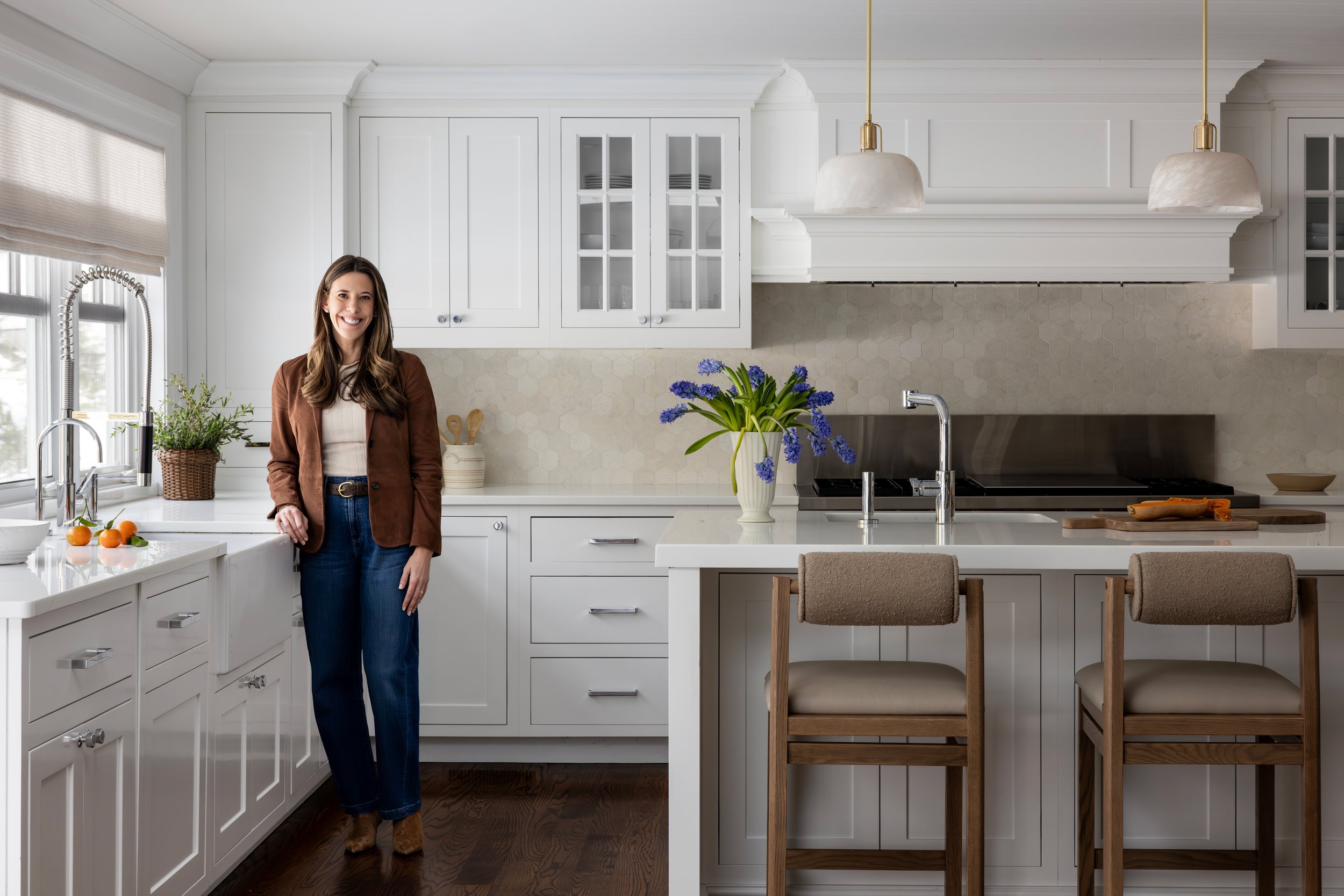 A woman standing in a bright, modern white kitchen with a vase of blue flowers on the island, chairs, and natural light coming from the window.