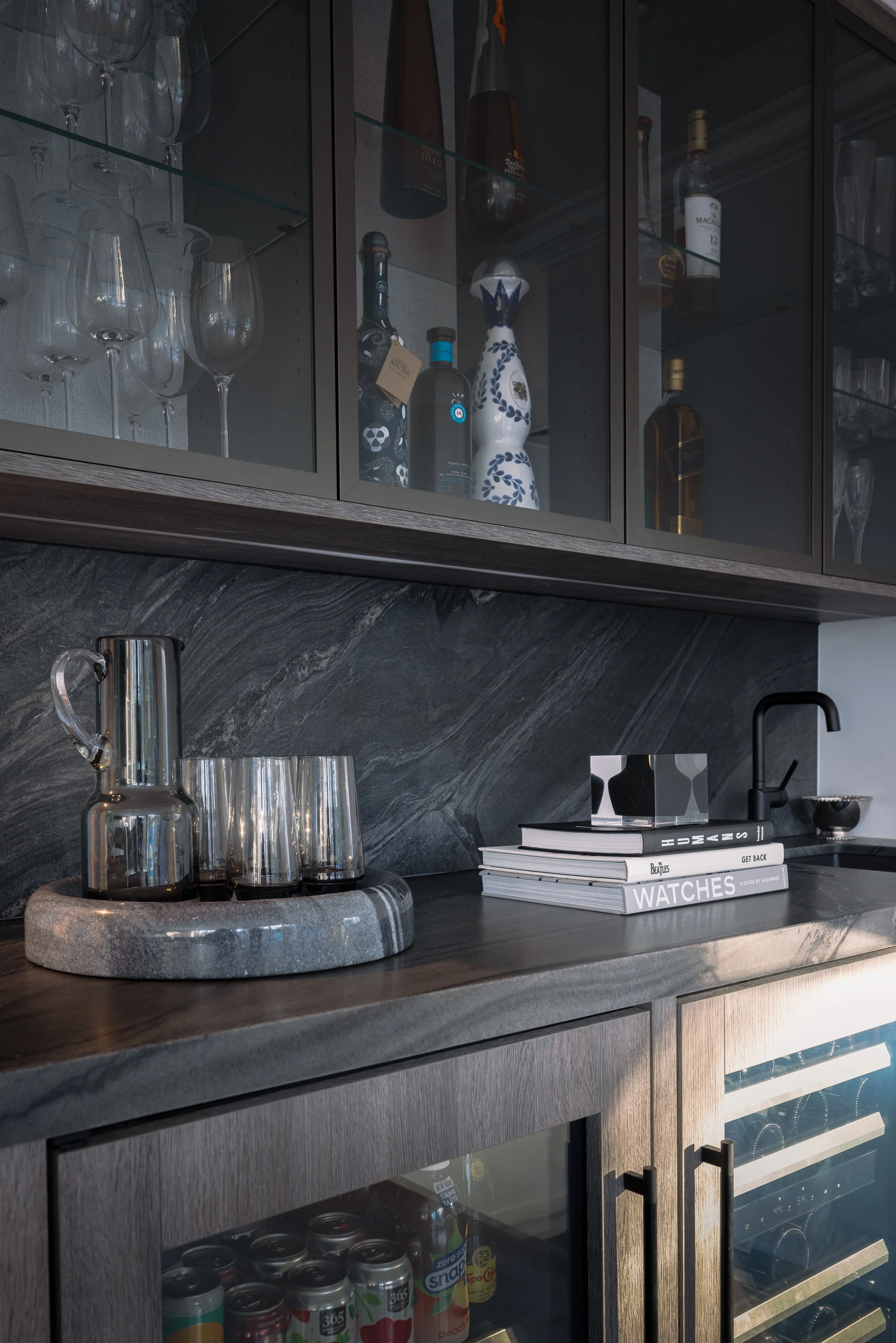 Kitchen counter with a stone tray holding a glass pitcher and three glasses, stacked books titled "Watches" and "Guides of HOGAN" beside a black faucet, dark wood cabinets with glass doors showing wine glasses, bottles, and decorative ceramics inside