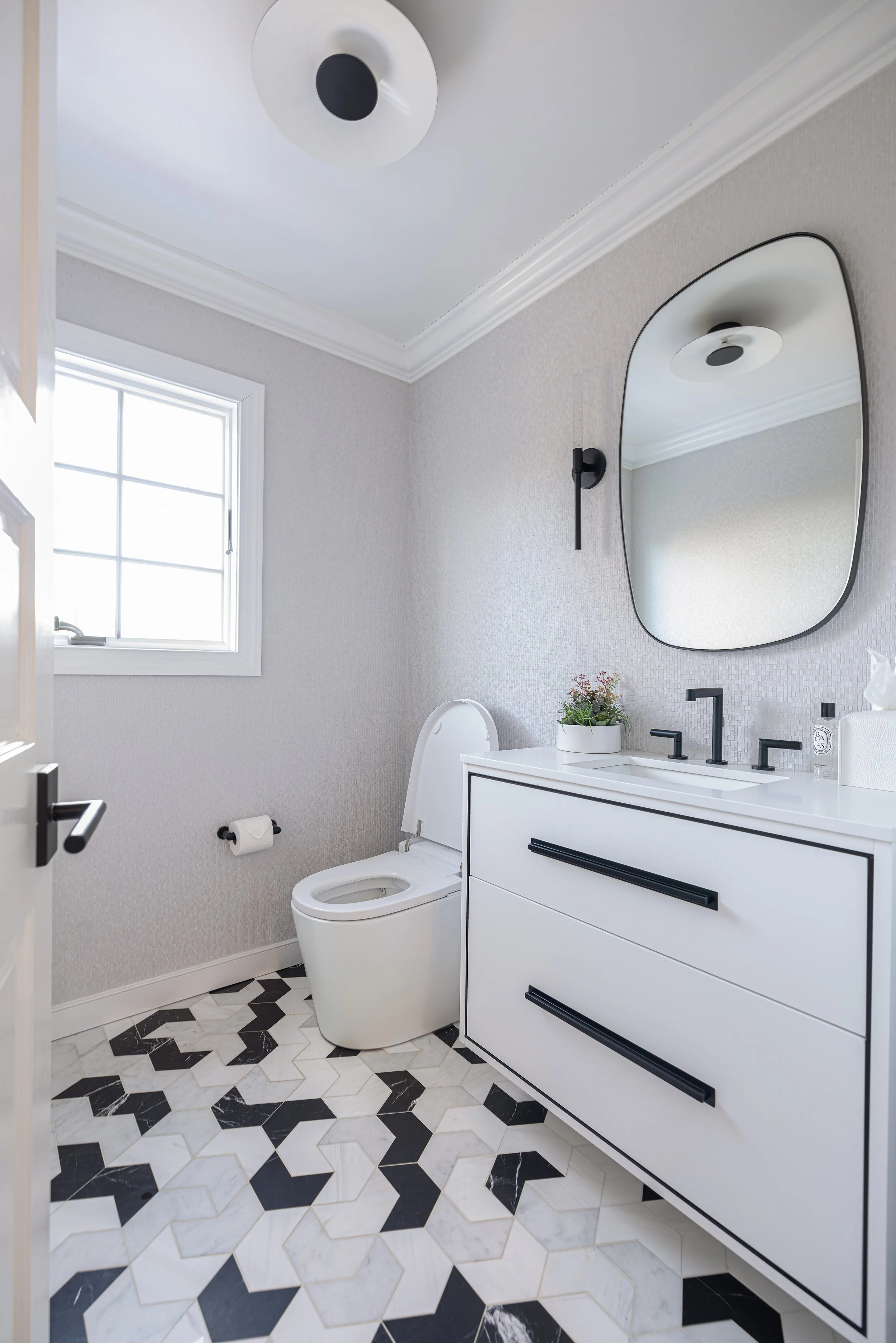 Modern bathroom with white toilet, white vanity with black handles, black faucet, oval mirror, black wall sconce, small plant, tissue box, and soap, with natural light from a window and geometric black and white floor tiles.