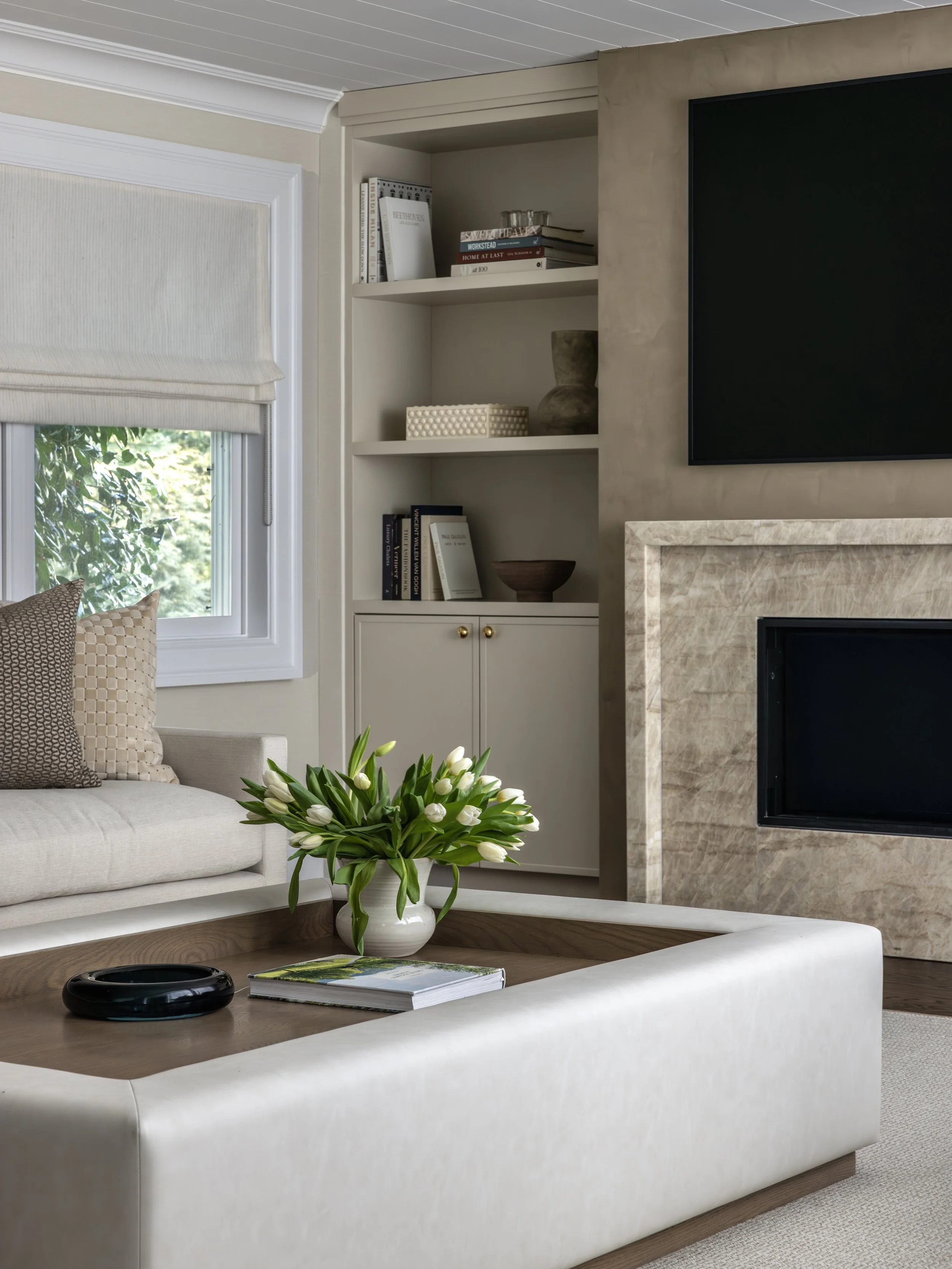 Cozy living room with a beige sofa, a wooden coffee table with a vase of white tulips, a fireplace with a stone surround, a built-in bookshelf with books and decorative items, and a window with a Roman shade.