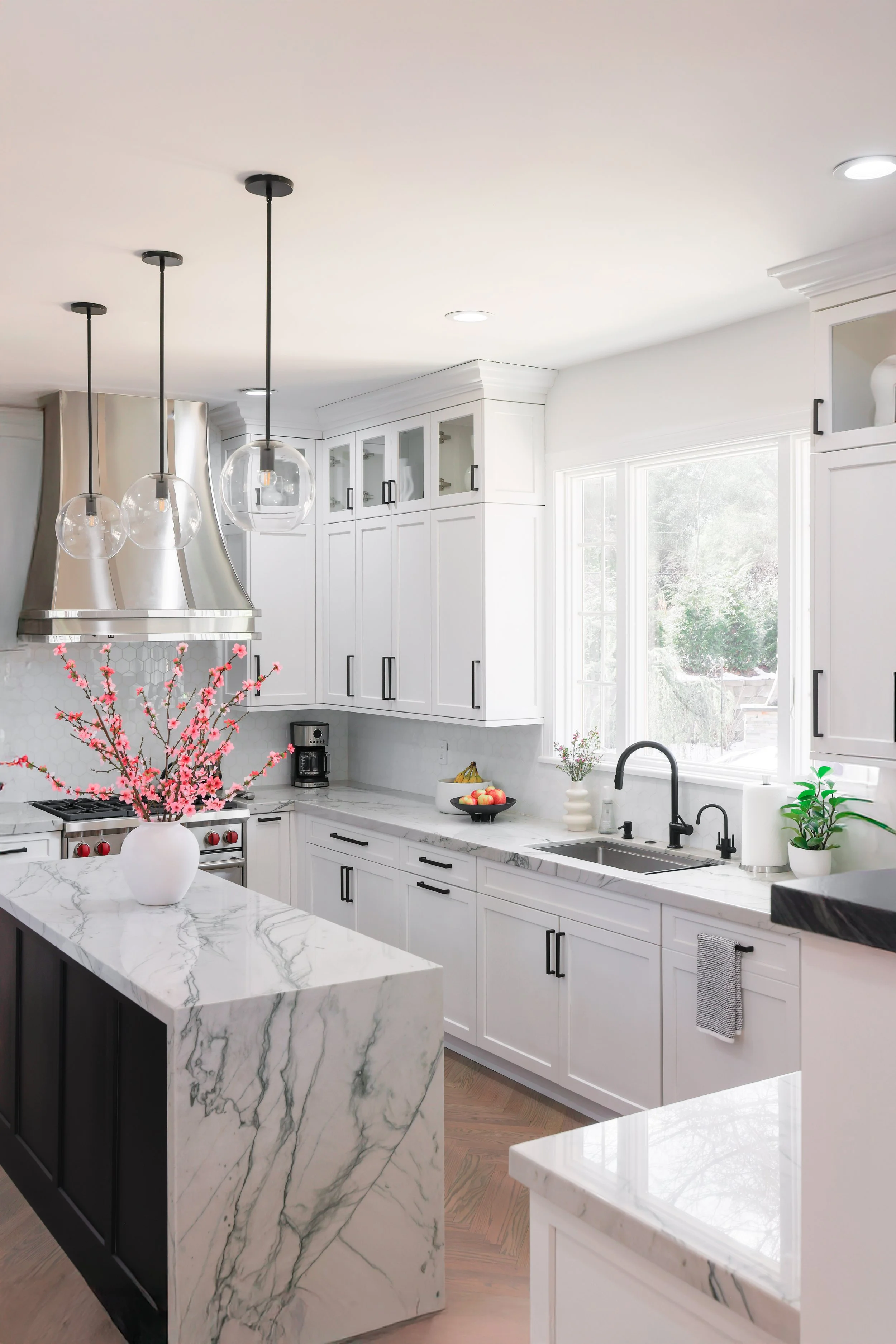 Bright modern kitchen with white cabinetry, black hardware, and marble countertops. A pink flowering plant in a white vase is on the island, with pendant lights hanging above.