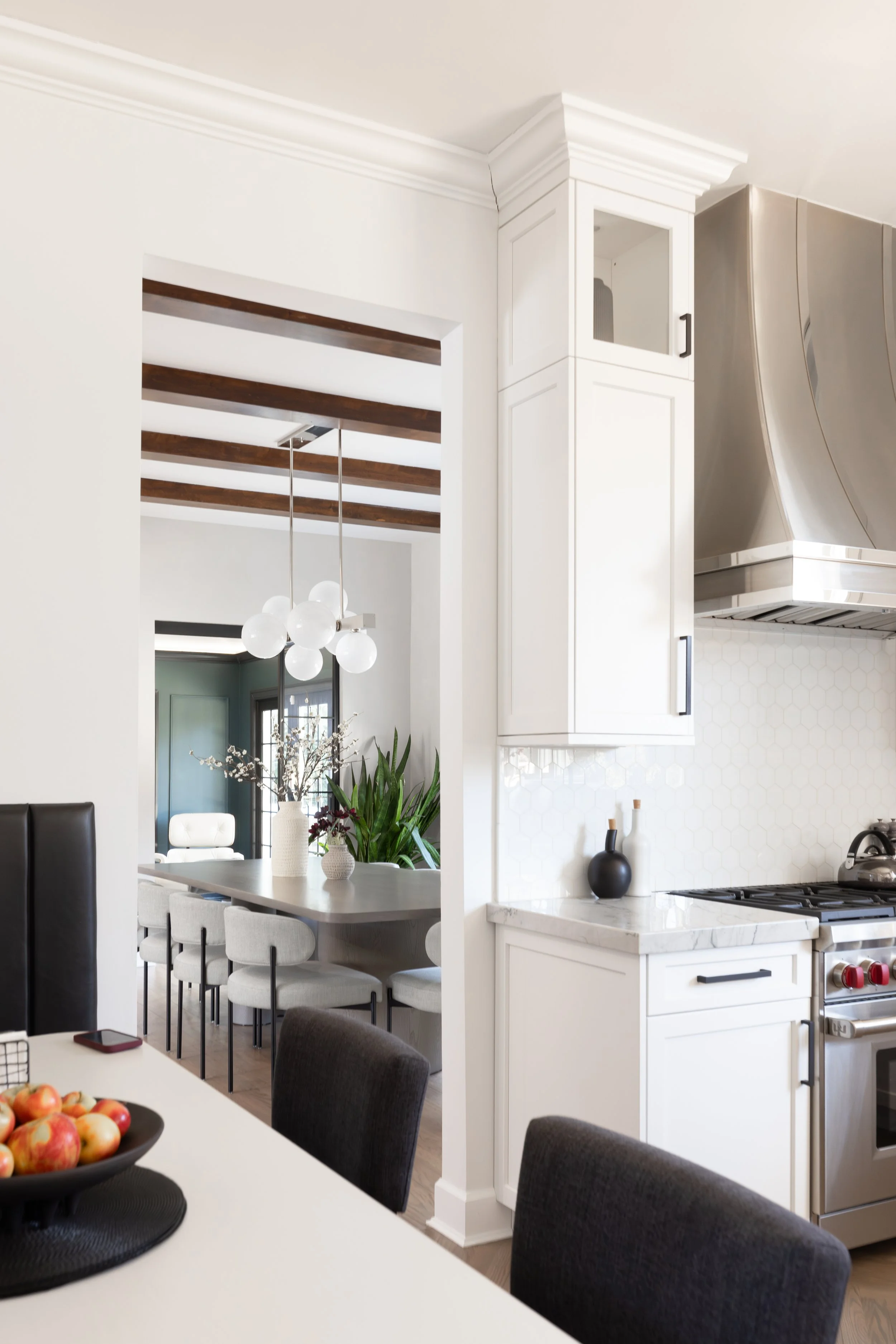 Modern kitchen with white cabinets, a stainless steel stove and range hood, a marble countertop, black vases, and a view into a dining area with a large table, cushioned chairs, a hanging chandelier, and a vase with flowers.