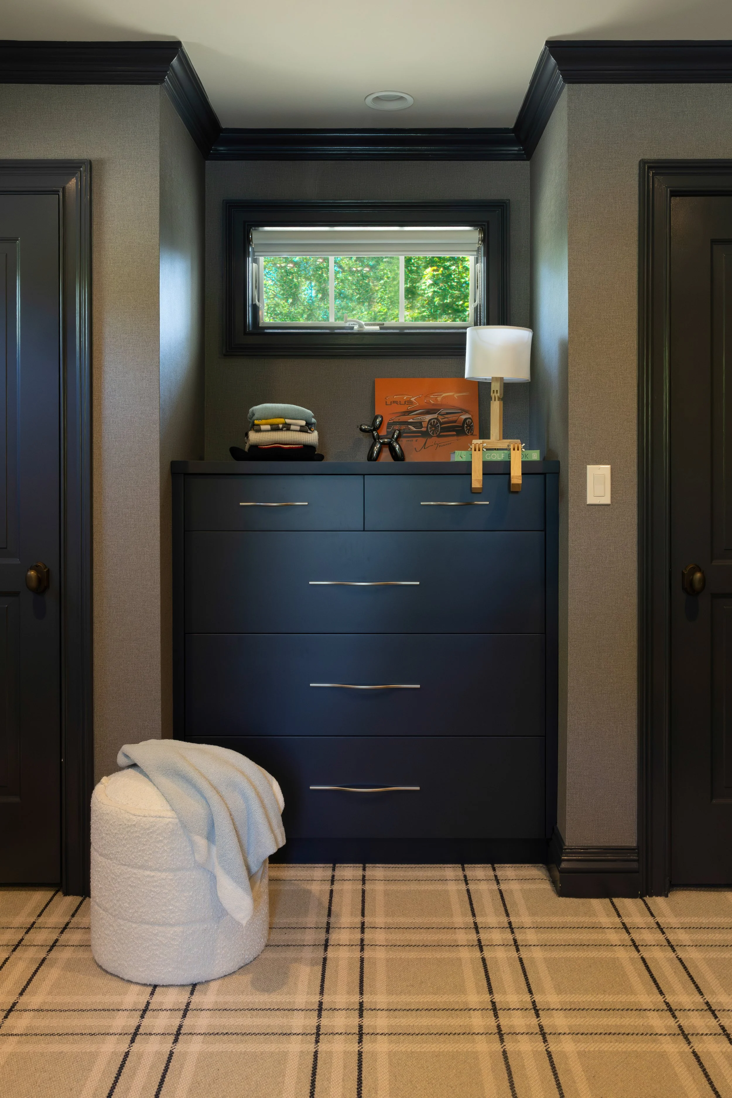 Dark blue dresser with six drawers, a small window above, a white lamp, a framed picture of a sports car, a black dog figurine, and a stack of folded clothes on top. A white textured ottoman with a blanket draped over it is on the beige checkered car