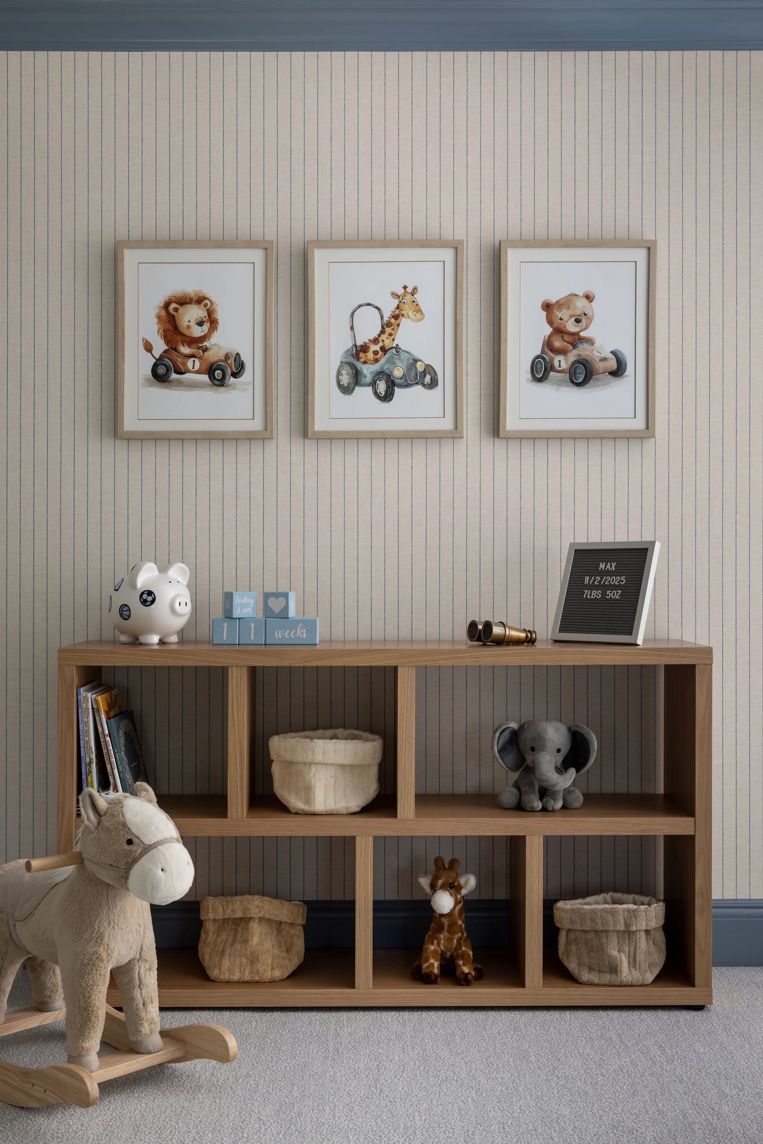Decorative nursery wall with three framed animal illustrations of a lion, giraffe, and bear in toy cars, above a wooden kid's bookshelf with toys, books, and a piggy bank, a plush horse on a wooden rocker, and a framed age and weight chart.