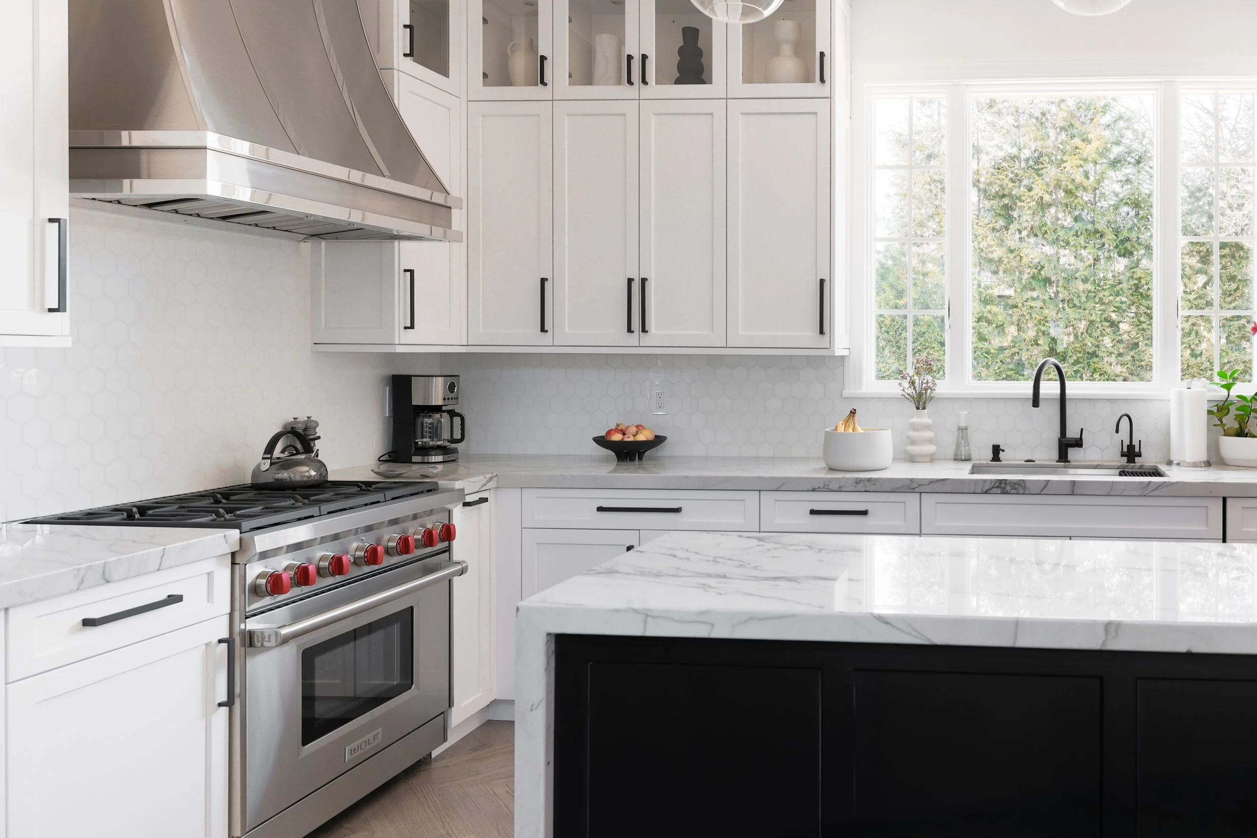 Modern white kitchen with marble countertops, black hardware, stainless steel stove, and large windows with view of trees.