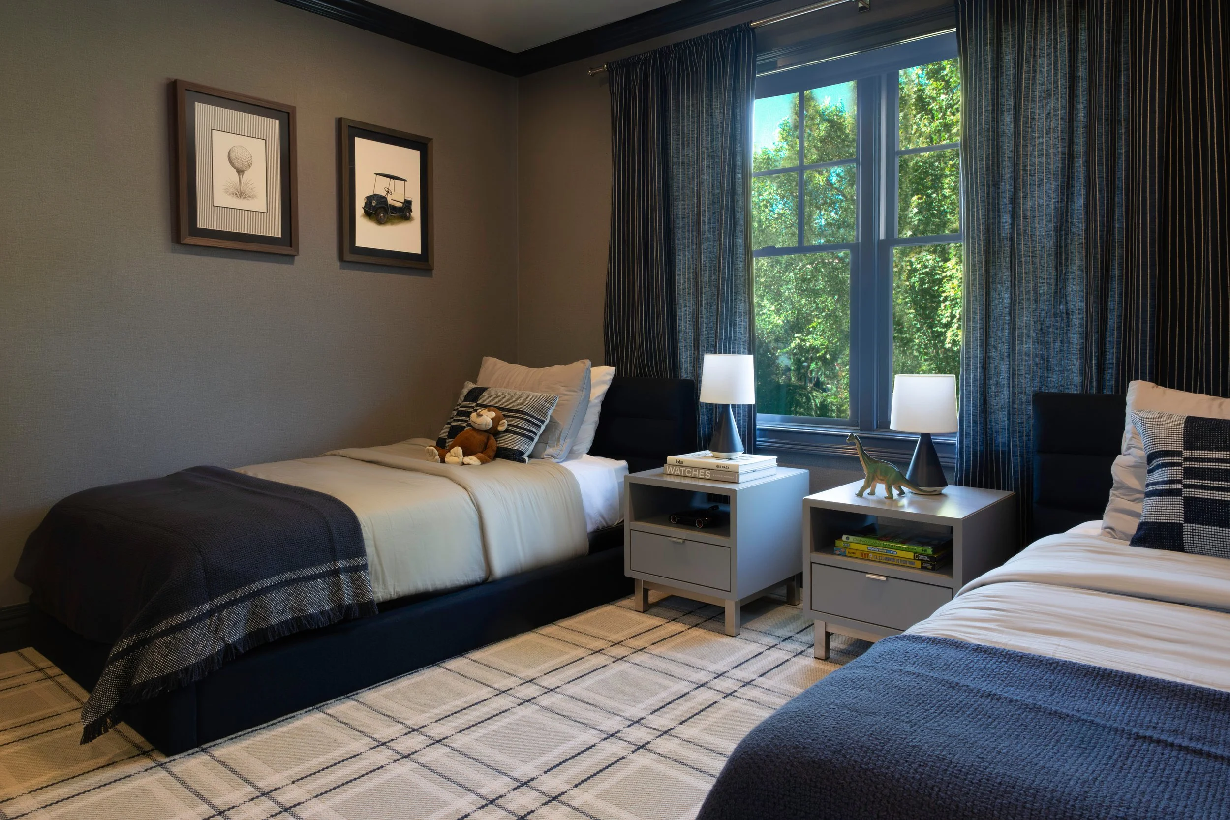 Bedroom with two beds, nightstands, lamps, window with blue curtains, framed pictures on wall, plush toys, and decorated with neutral tones.