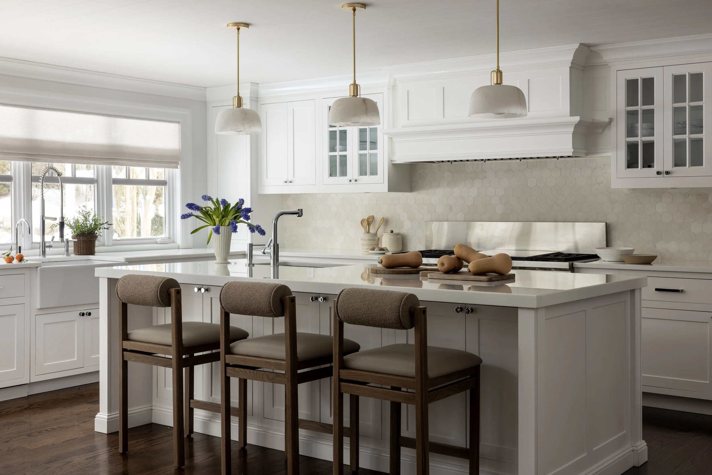 Bright white kitchen with large island, beige chairs, wooden cutting boards with gourds, blue flowers in a white vase, window with blinds, and pendant lights.