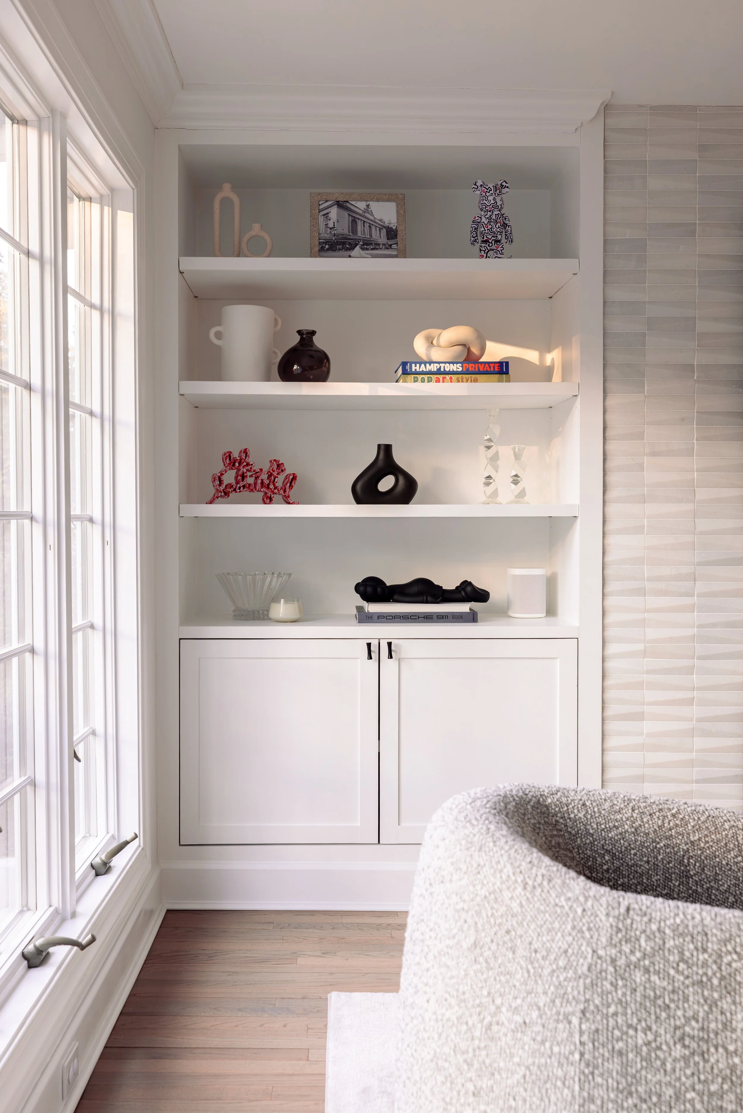 A built-in white bookshelf with decorative items and books, next to a window with sunlight and a beige textured upholstered chair in the foreground.