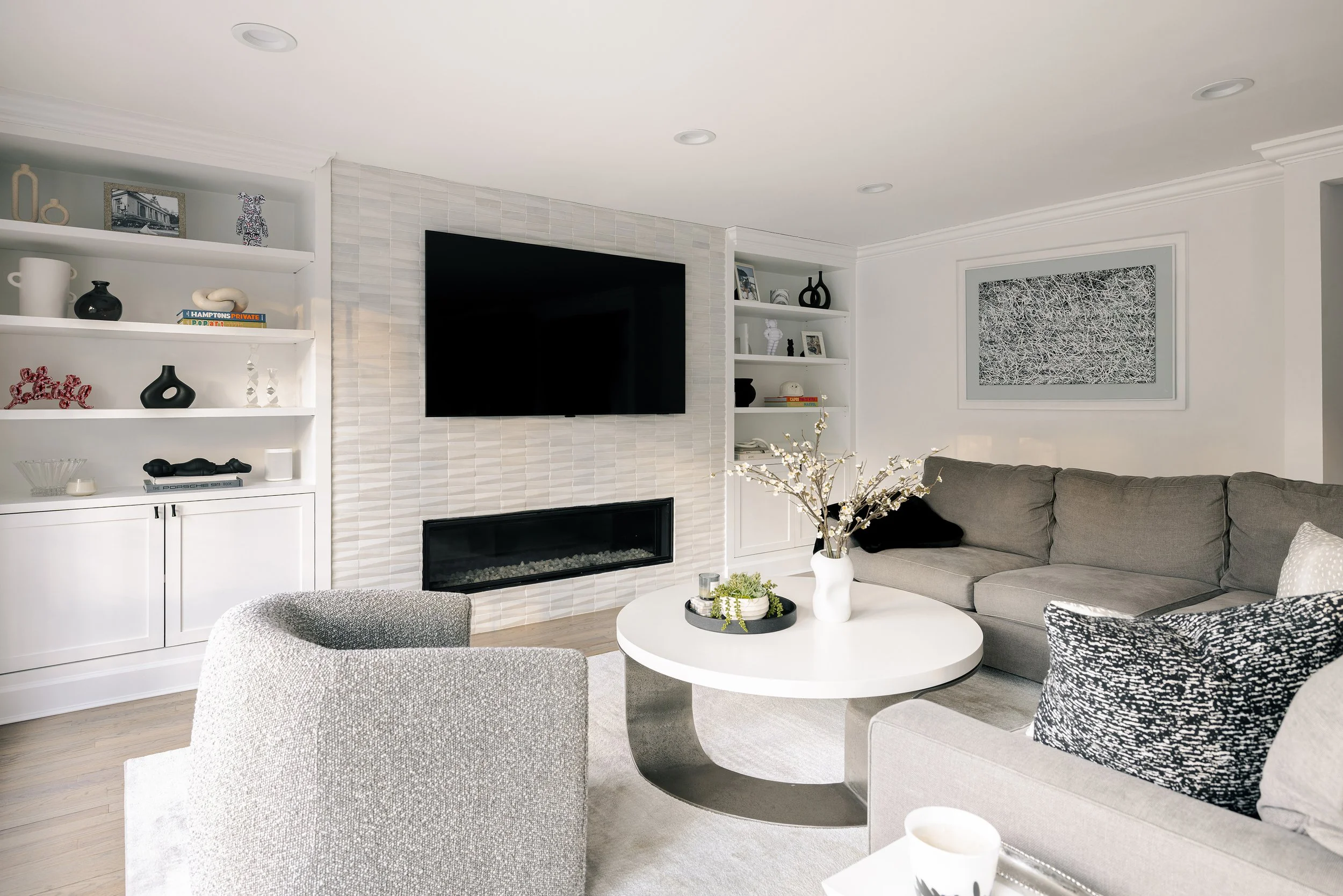 Modern living room with white shelving, a wall-mounted TV, a fireplace, gray sectional sofa with black and patterned pillows, a round white coffee table with decorative items and a vase with branches, and contemporary art on the white walls.