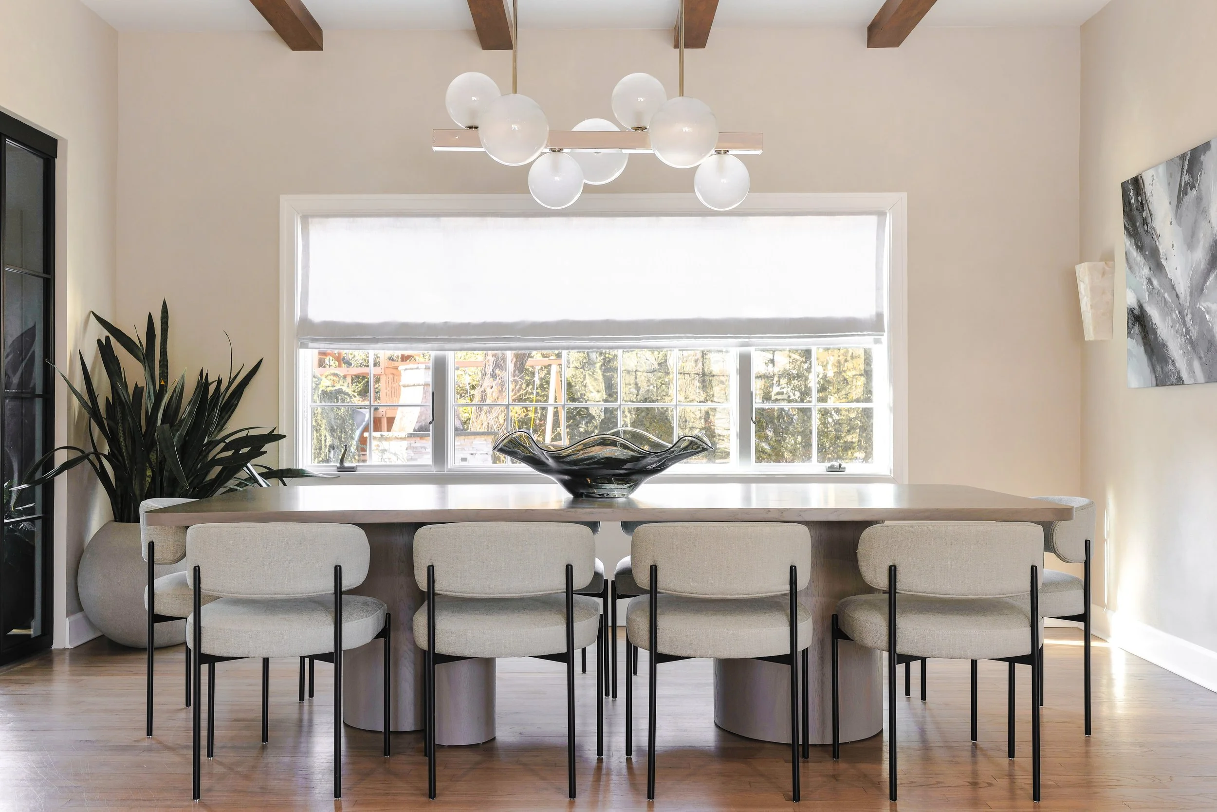 A modern dining room with a large wooden table and six upholstered chairs with black metal legs. A black decorative bowl is on the table. There are large windows with a white roller shade, and a contemporary chandelier with multiple white globes hang