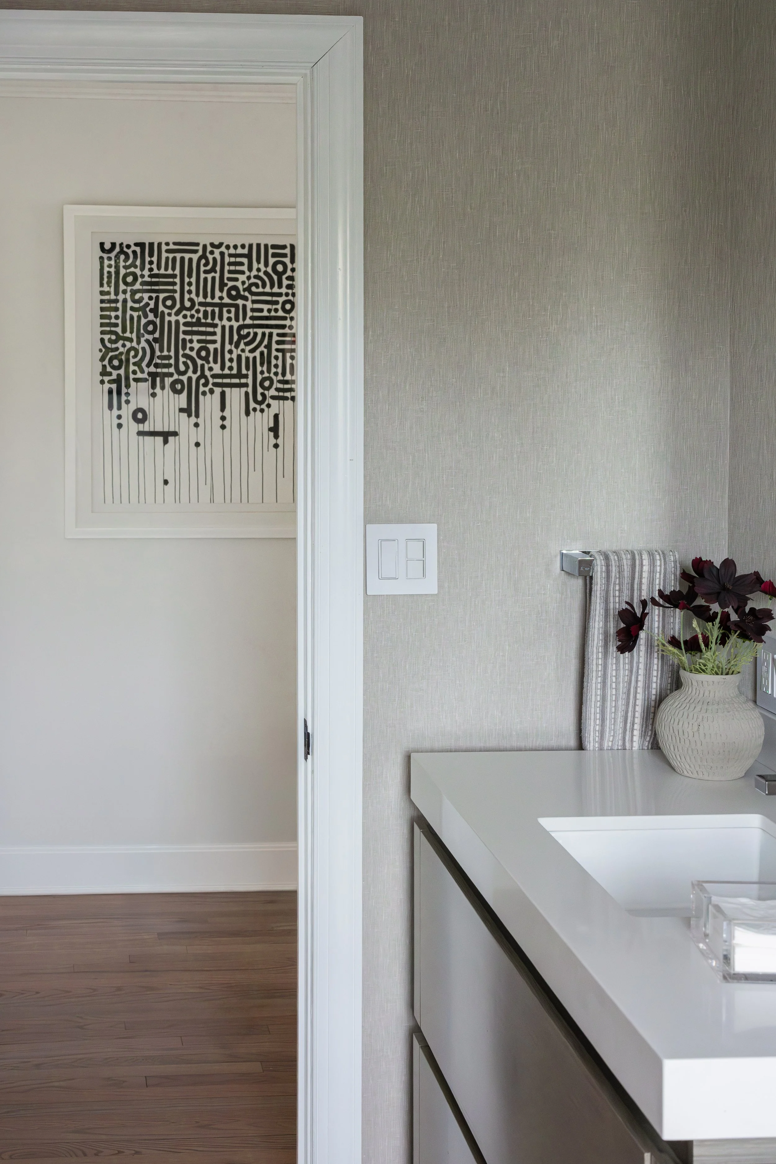 Part of a modern bathroom with a white countertop, vase with dark purple flowers, and a towel hanging on a rack, with a gray textured wall and a light switch.