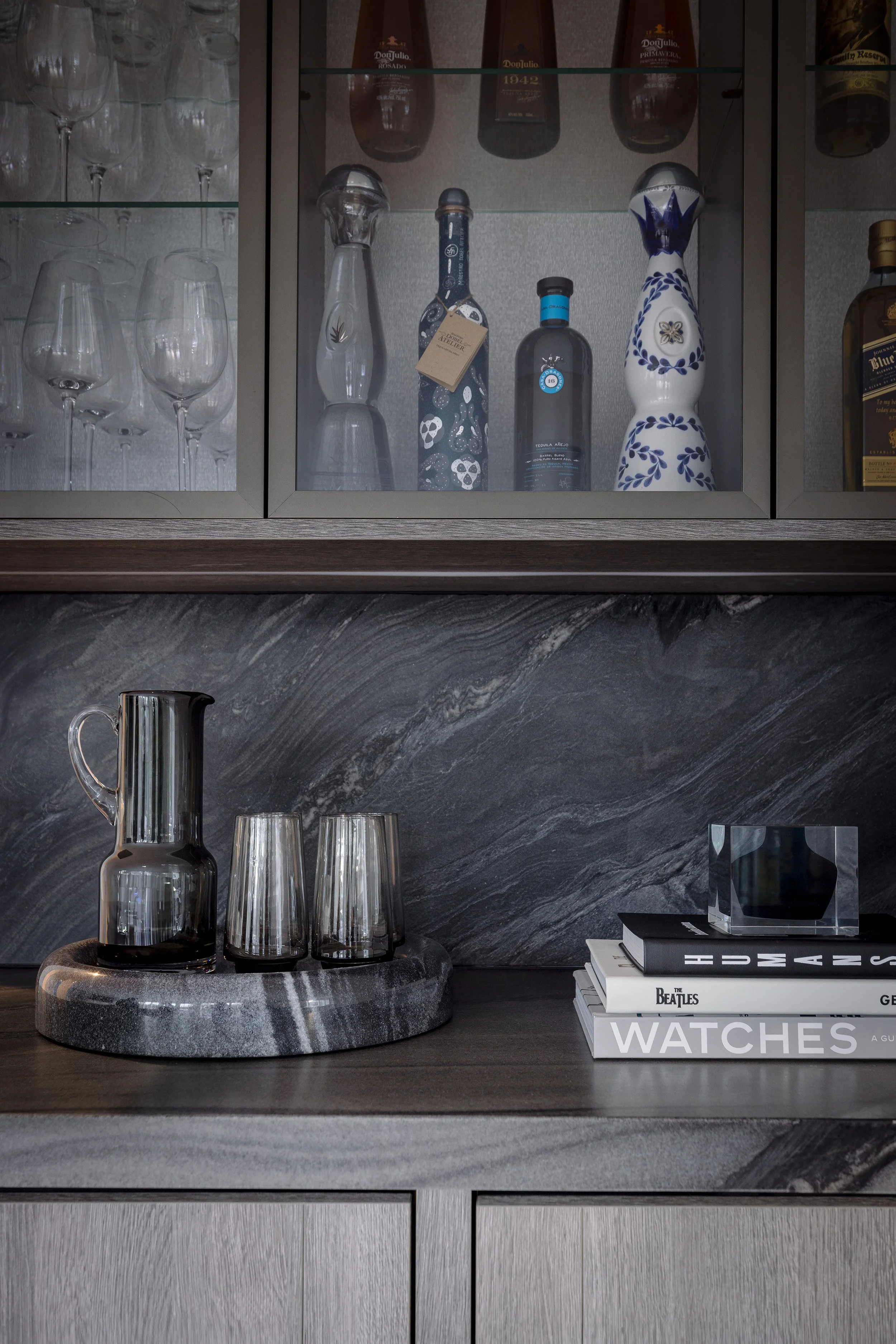 A bar area with glassware, liquor bottles, a tray with glasses, books, and decorative items set against a dark marble backsplash.