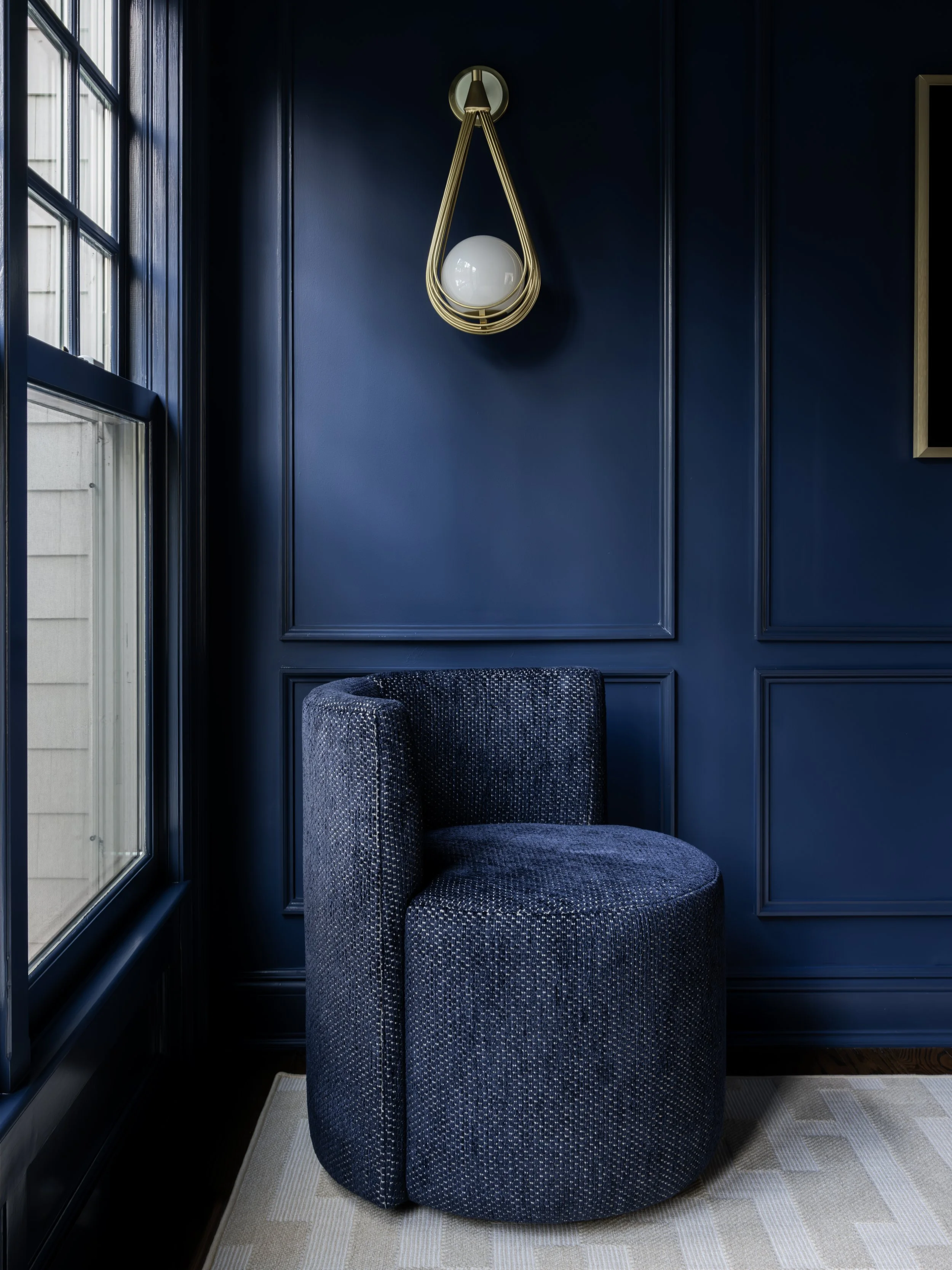 Blue upholstered armchair in a corner next to a window with navy blue walls and decorative wall paneling, with a modern brass wall sconce above.