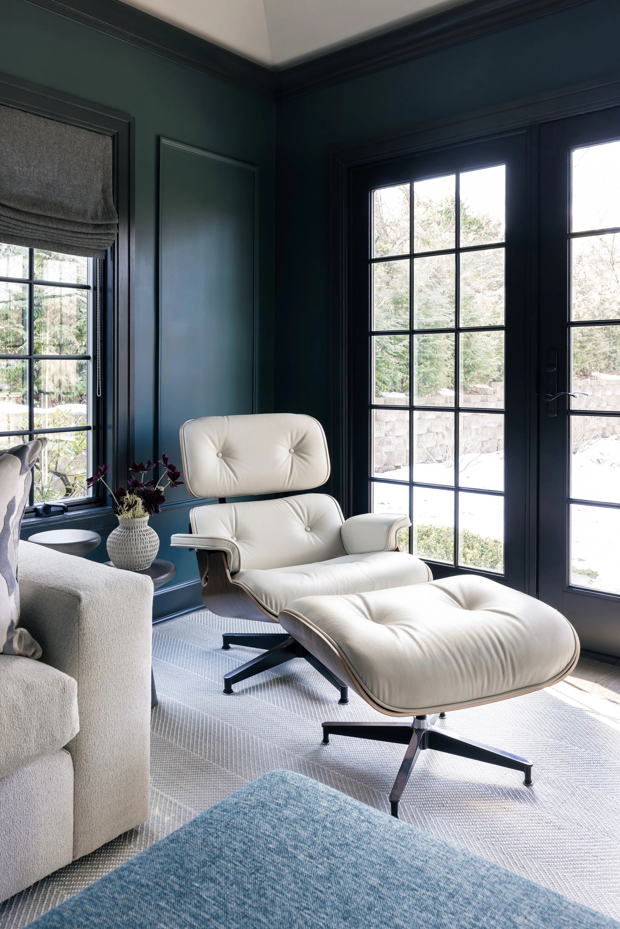 A modern living room corner with a white lounge chair and matching ottoman, a side table with a white vase and dark purple flowers,, a beige sofa, large glass doors, and a window showing a snowy outdoor scene.