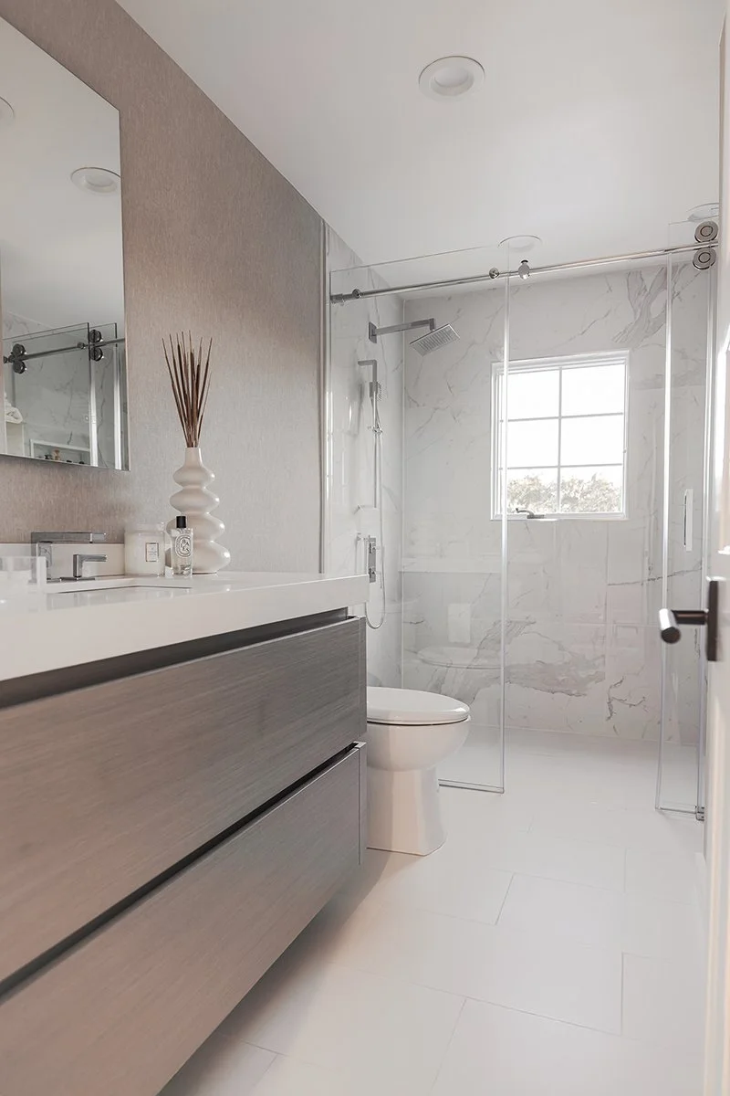 Modern bathroom with a vanity, a sink, and a mirror on the left, decorative items on the vanity, a toilet, and a walk-in shower with glass doors and a window on the far wall.